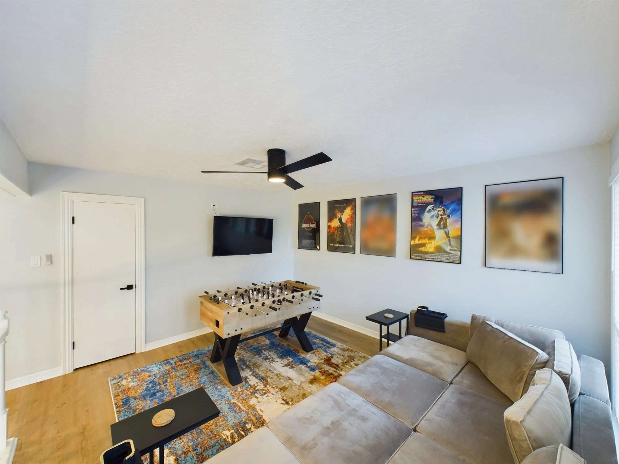Living room with beige couch, foosball table, black TV mounted on white wall, posters, ceiling fan, small side table, and a colorful area rug.