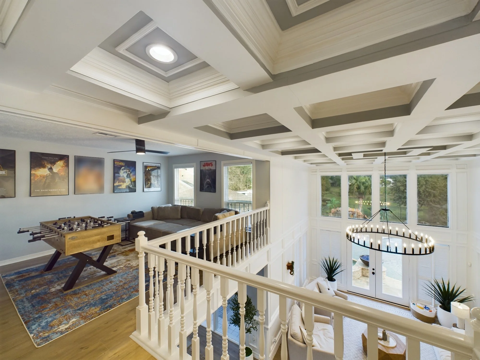 A multi-level living room with a white staircase, large windows, and a ceiling featuring a geometric tray design with a ceiling light. The room has a tan sectional sofa, a foosball table, framed posters on the wall, and a decorative chandelier hangin