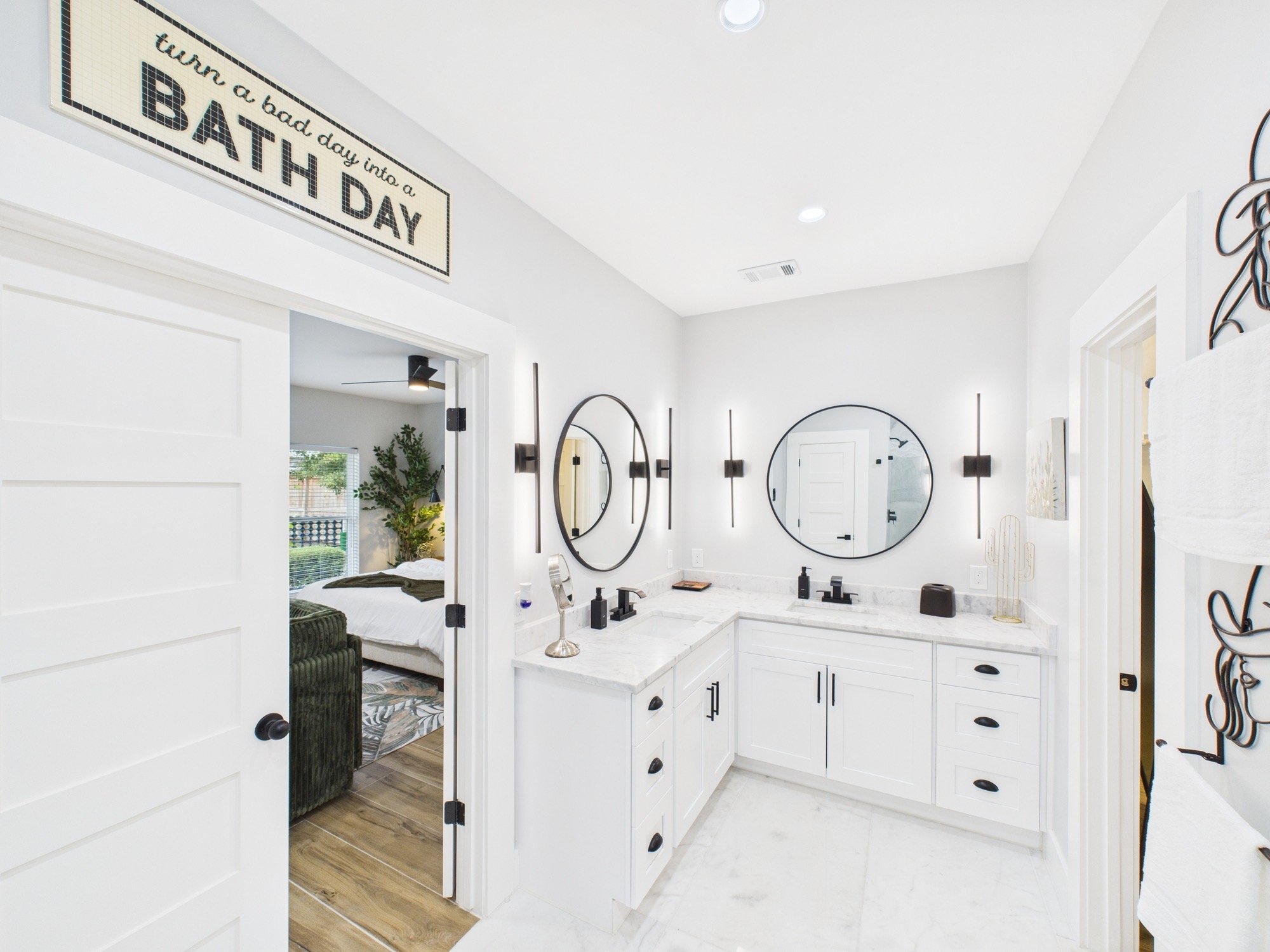 Bathroom with white cabinets, black hardware, oval mirrors, and modern wall lights, viewed from doorway. The room is bright with white walls and marble floors.