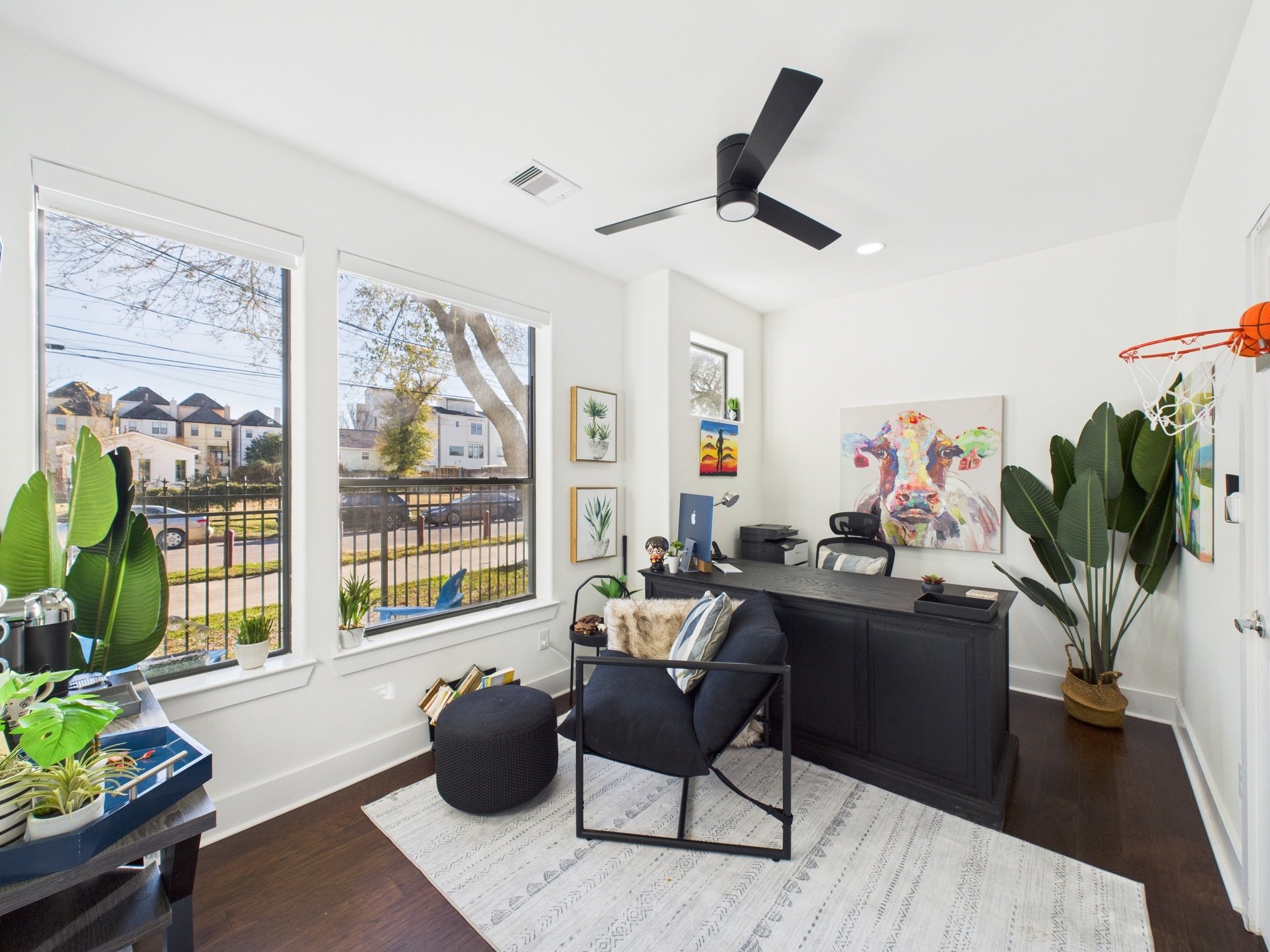 Bright and modern home office with large windows, potted plants, contemporary art, a black desk, black chair, and ceiling fan.