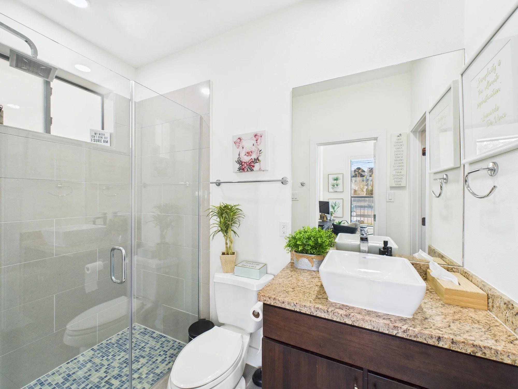 A modern bathroom with a shower area, white double sinks, mirror, and decorative wall art with a pig painting and various framed pictures. There are green plants and a granite countertop.