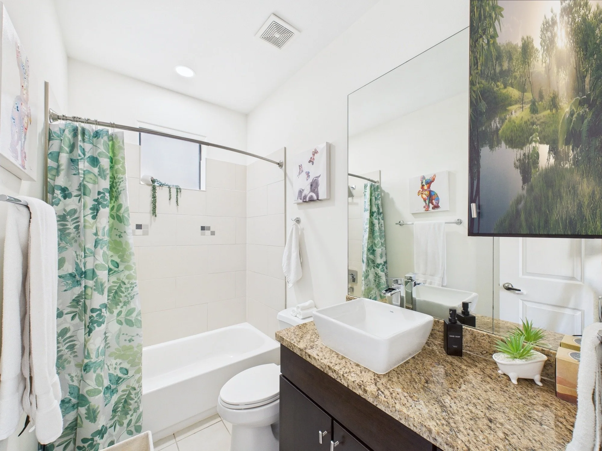 A bathroom with a white bathtub and shower curtain with green leaf pattern, a white toilet, a granite countertop with two white vessel sinks, a large mirror, artwork of a rabbit and a dog, and decorative plants.