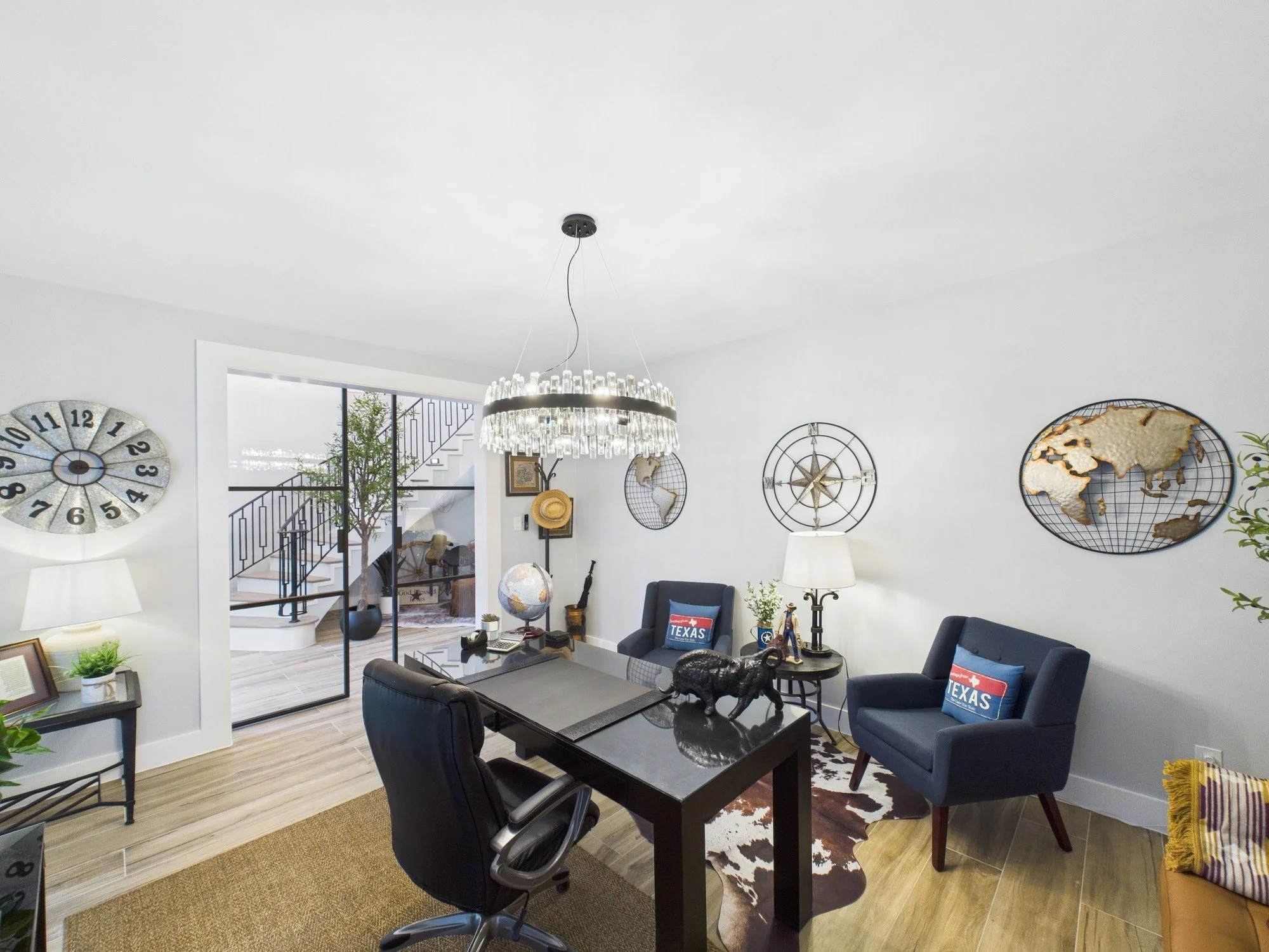 Living room with black dining table, six black chairs, blue armchairs with Texas pillows, decorative world map wall art, globe, plants, lamps, and staircase visible through doorway.
