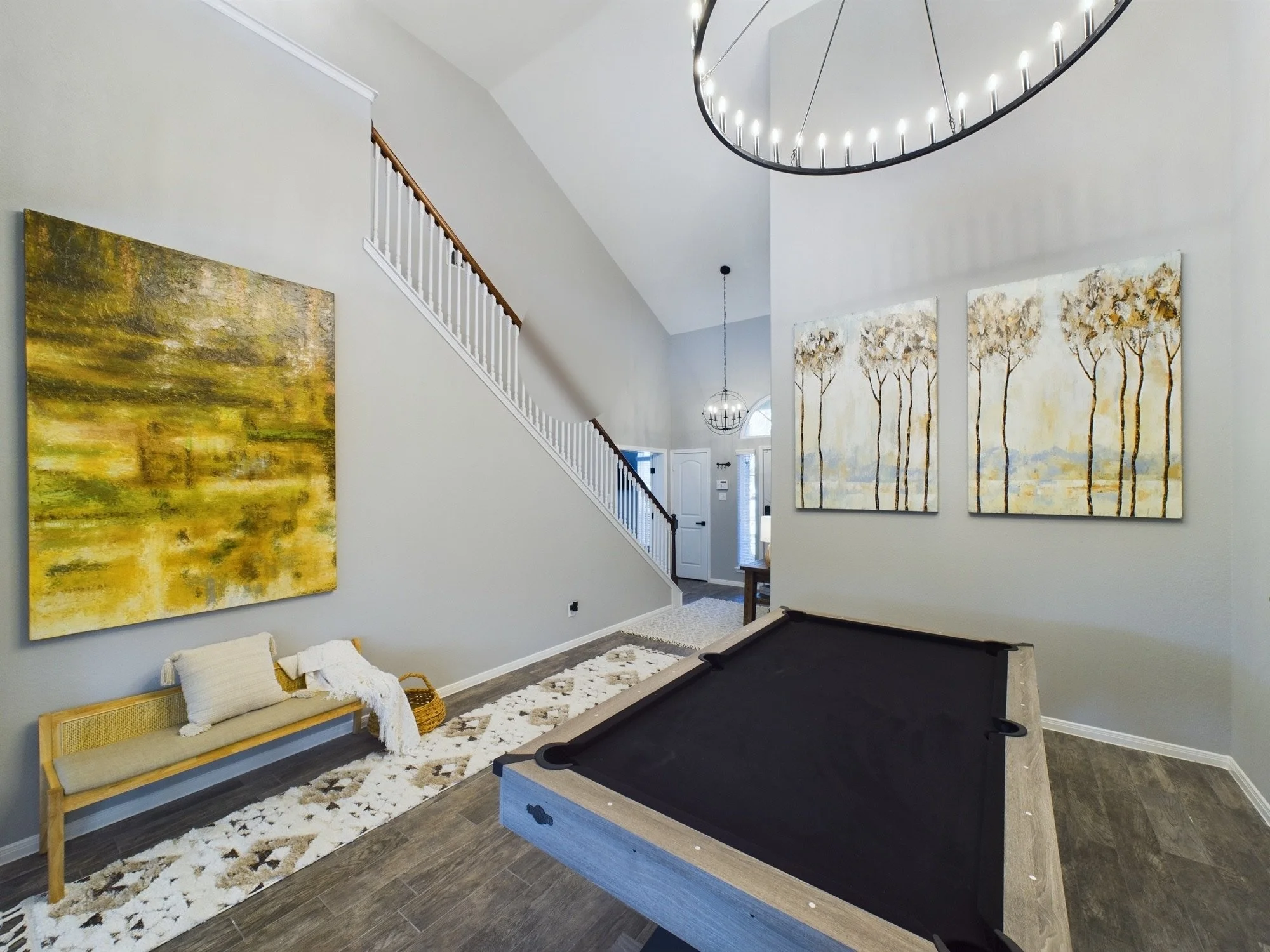 A living room with a staircase and high ceilings, decorated with three large abstract paintings of trees. There is a black pool table, a wooden bench with cushions, and a white and beige rug on dark hardwood floors. The room has hanging light fixture