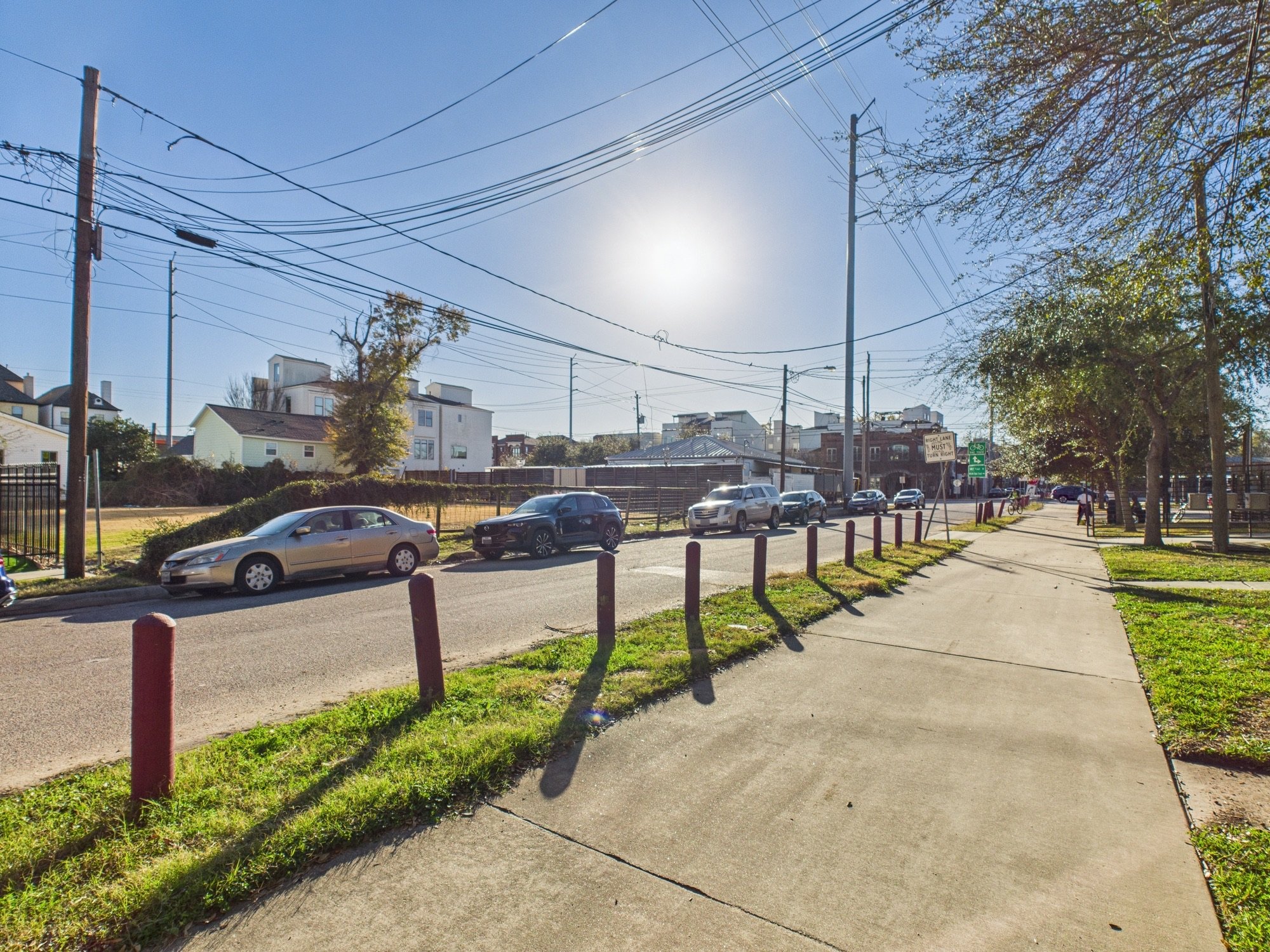 A sunny urban street scene with parked cars, trees, power lines, and residential buildings under a clear blue sky.