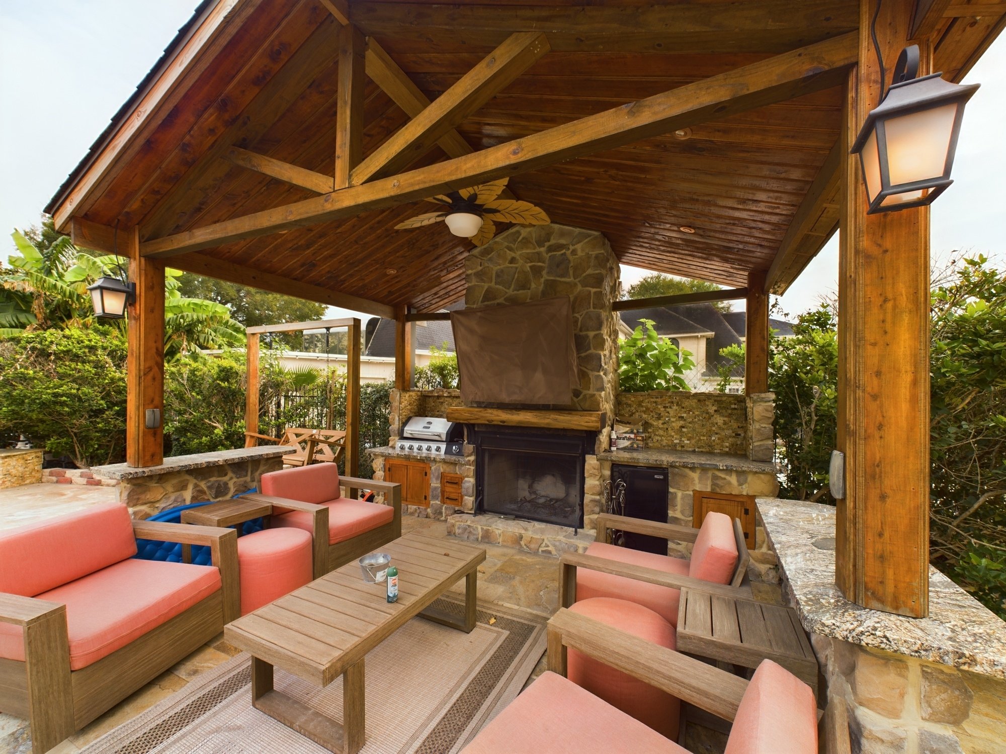 A covered outdoor patio area with a wooden roof and stone fireplace. The space is furnished with pink cushioned chairs and wooden tables, and is decorated with two wall-mounted lanterns and a ceiling fan with leaf-shaped blades. Green foliage surroun
