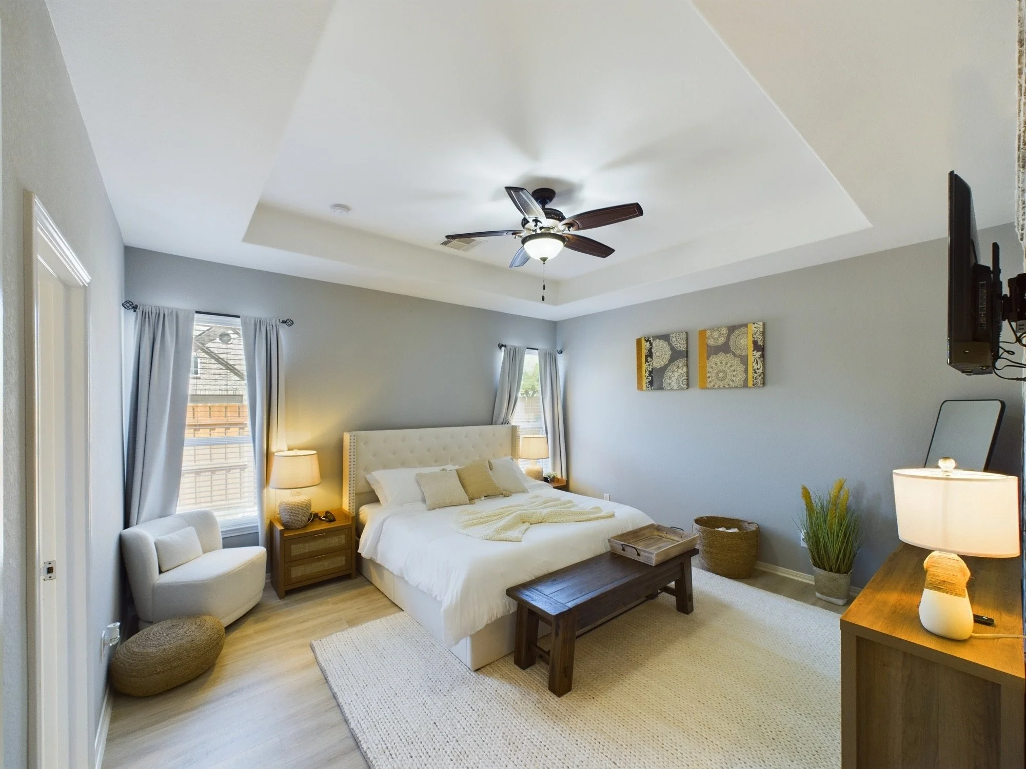A cozy bedroom with a white bed, beige headboard, and wooden furniture. There are two bedside tables with lamps and a small white chair in the corner. The room has light gray walls, two windows with white curtains, and a ceiling fan.