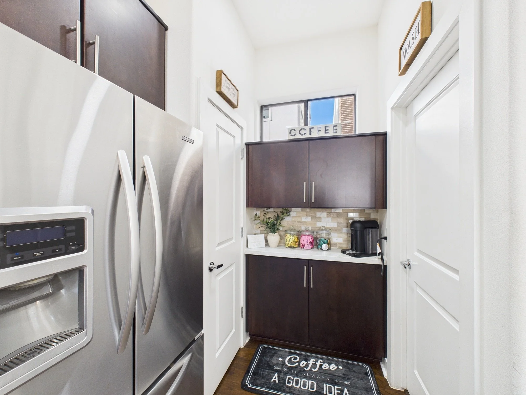 A small kitchen area with white walls and dark wood cabinets. There is a stainless steel refrigerator on the left and a coffee station on a white countertop with jars and a coffee machine. A black rug with white text reading, 'Coffee is always a good