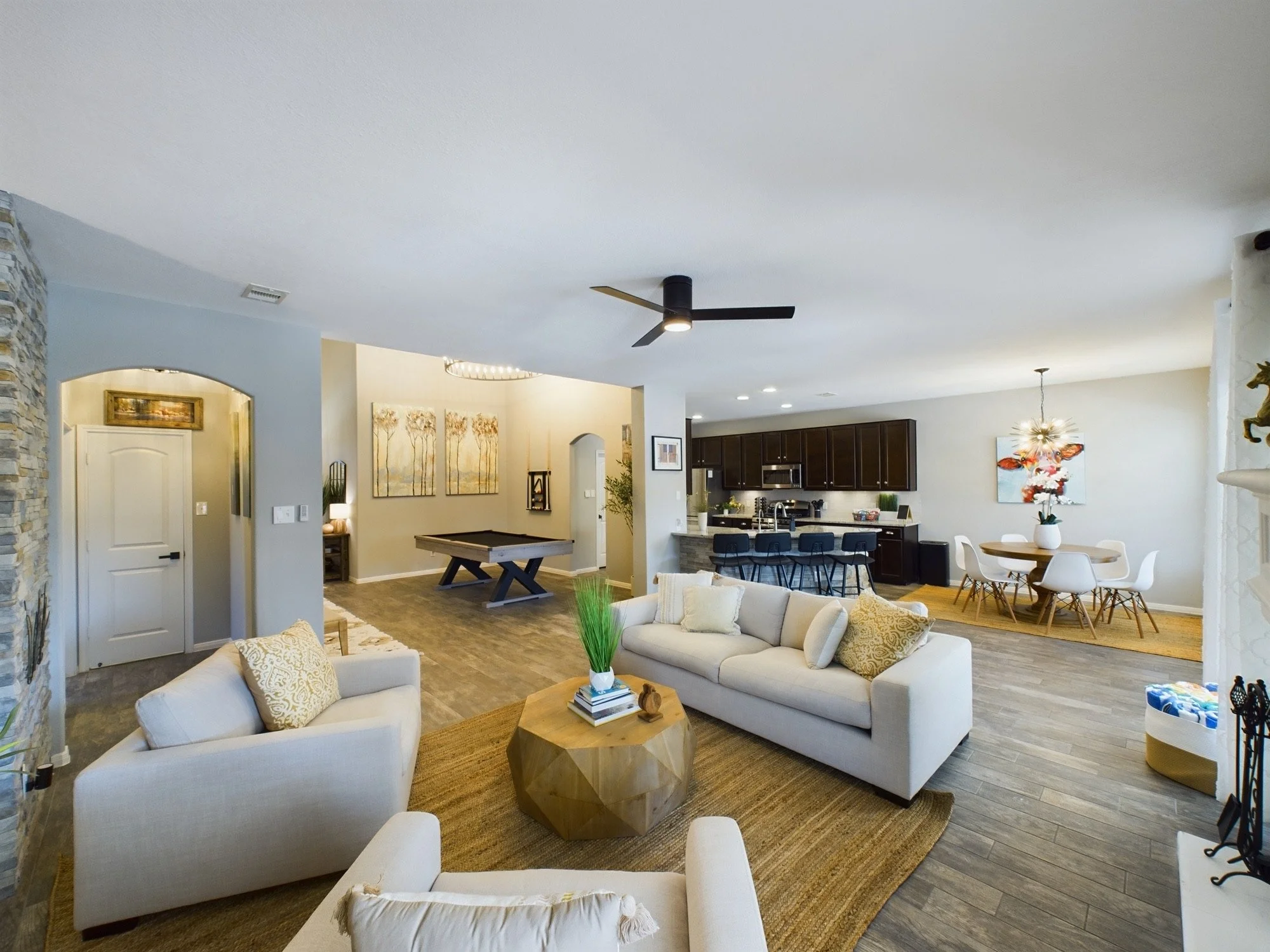 Open-concept living room and kitchen with modern decor, white sofas, wooden tables, artwork on walls, ceiling fan, and hardwood flooring.