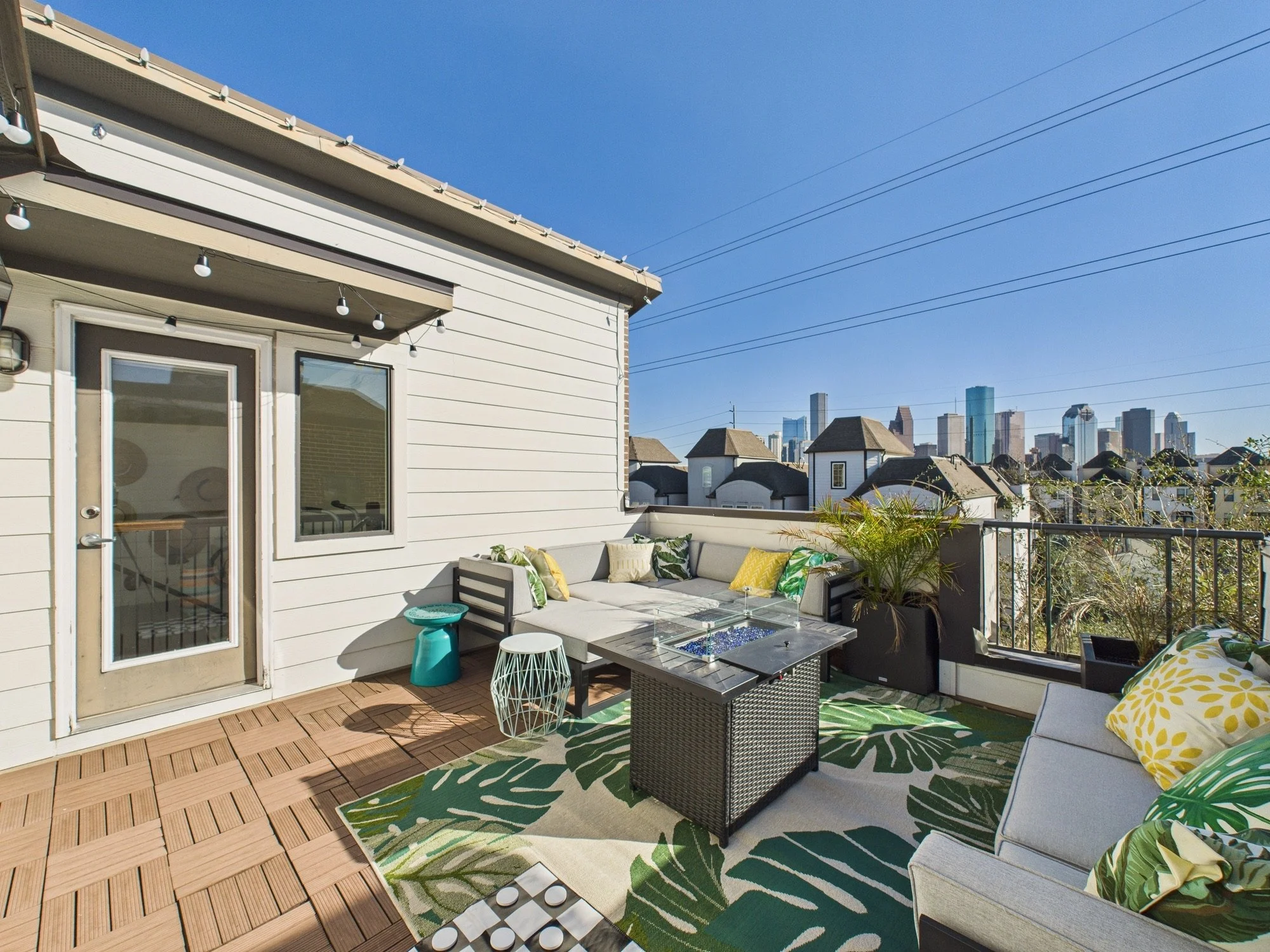 Urban rooftop patio with outdoor seating, potted plants, patterned rugs, and city skyline in the background.
