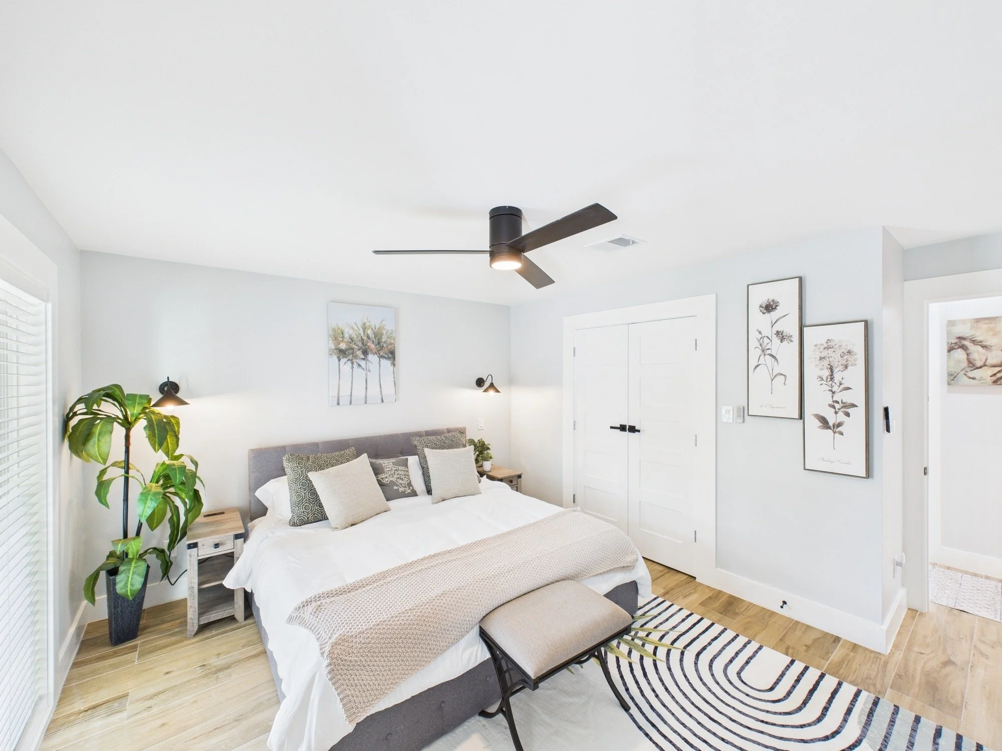 Bright bedroom with white walls, a large bed with white and beige bedding and multiple pillows, a black ceiling fan, artwork of flowers on the walls, a potted plant, and a patterned rug.