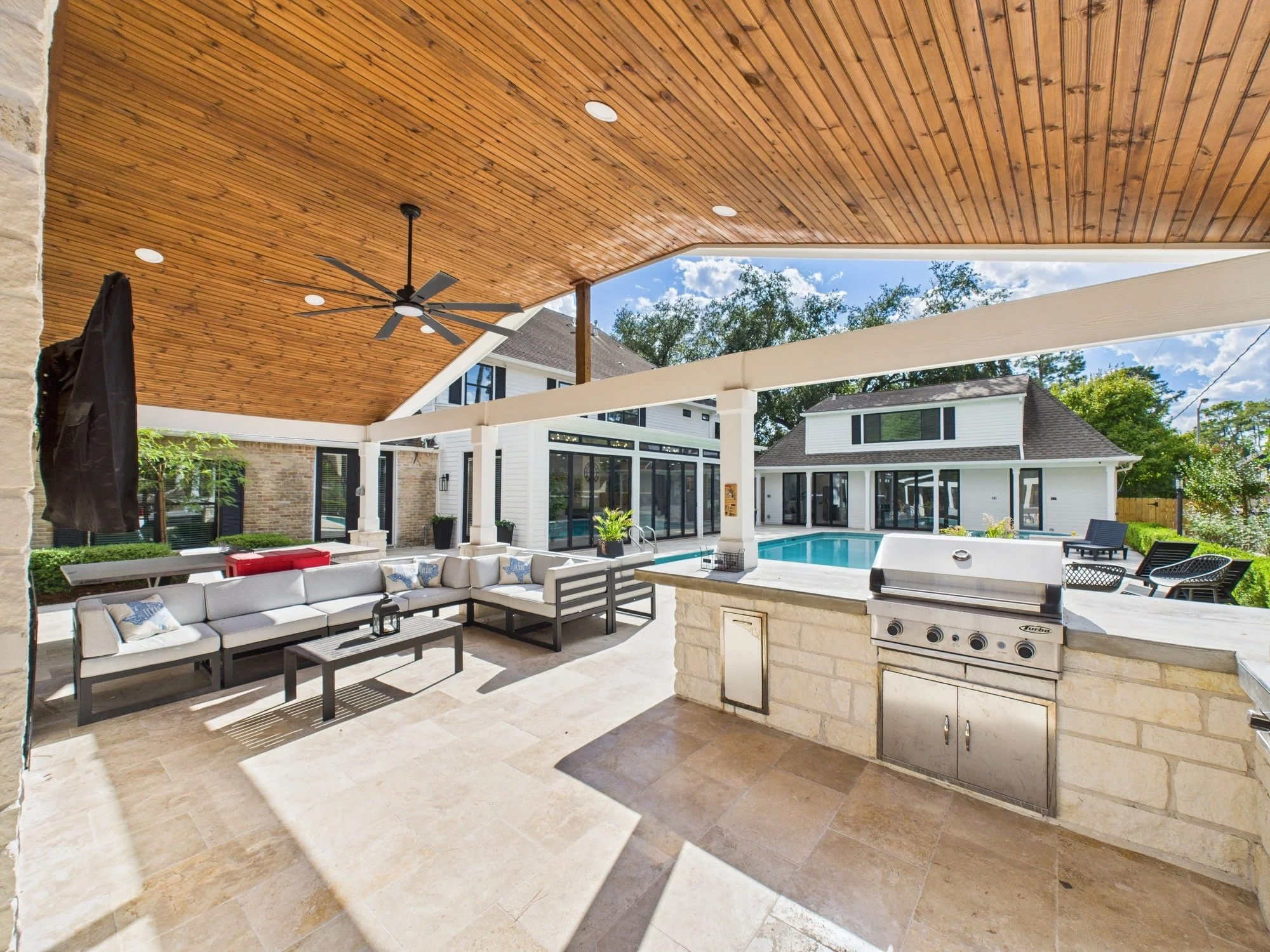 A covered outdoor patio with beige tiled flooring, built-in grill, and outdoor seating area. The ceiling of the patio is made of wooden planks with recessed lighting and a ceiling fan. In the background, there is a swimming pool and a white house wit