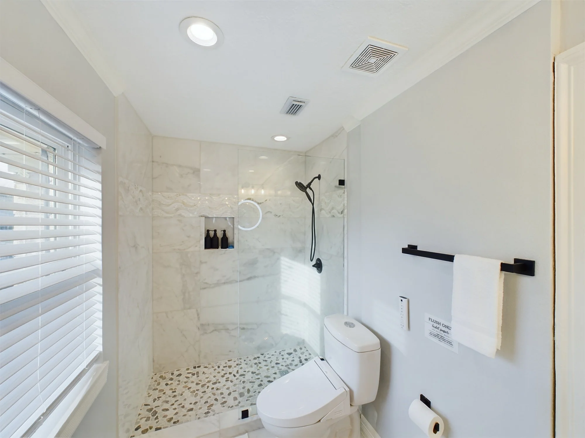 Modern bathroom with a shower area featuring marble tiles, a rain shower head, a glass partition, and a window with white blinds. The bathroom also contains a white toilet and a paper towel or tissue holder on the wall.