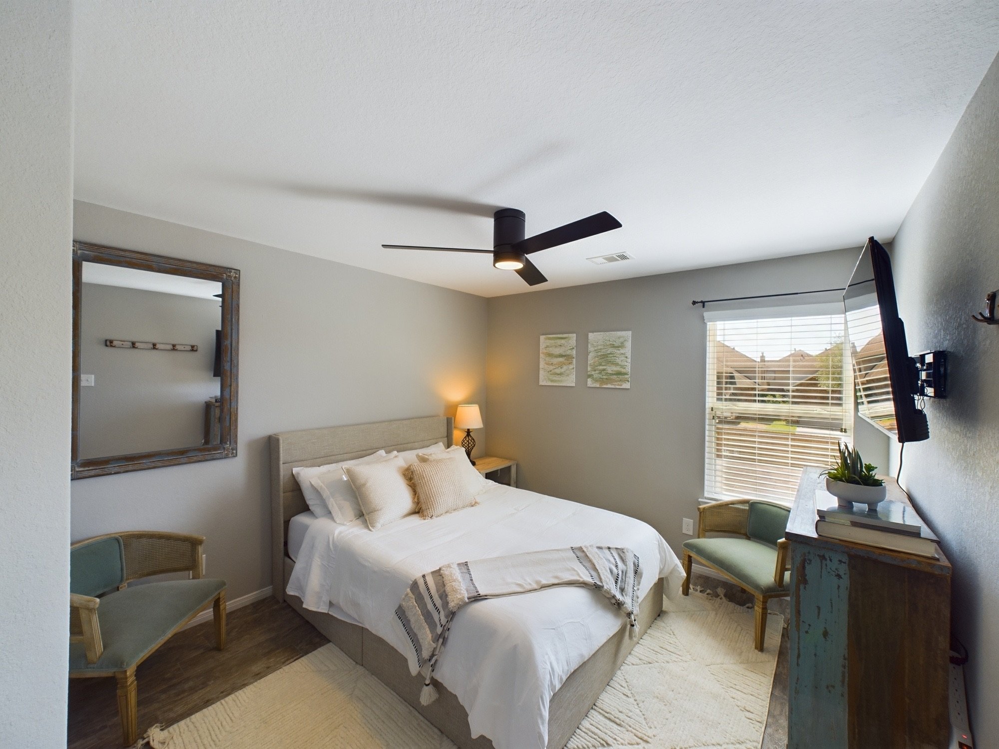 A bedroom with a bed, window with blinds, wall-mounted television, ceiling fan, desk, and chairs.