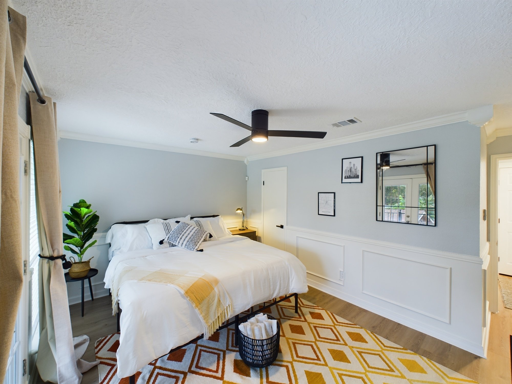 A bedroom with a bed covered in white bedding and decorative pillows, a nightstand with a lamp, a potted plant, framed art on the wall, a large mirror, a window with curtains, and a ceiling fan.