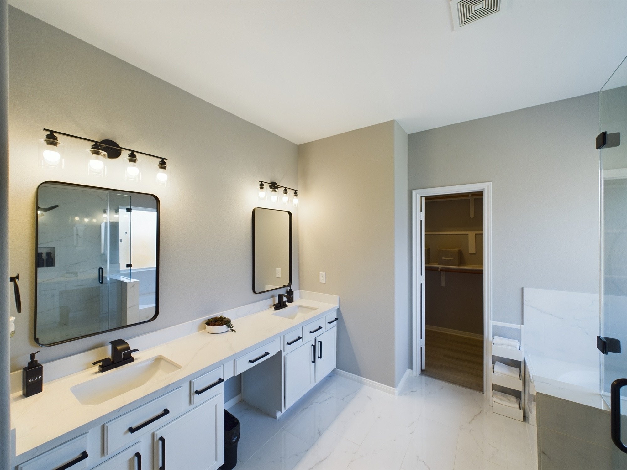 Modern bathroom with double vanity, black hardware, large mirrors, wall-mounted light fixtures, small potted plant, and open closet with shelves.