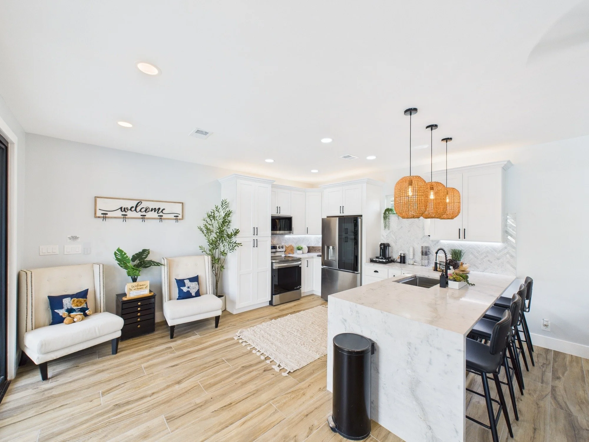 Bright, modern kitchen with white cabinets, stainless steel appliances, a marble island, and hanging wicker pendant lights. There is a welcome sign on the wall, two white chairs with decorative pillows, green plants, and a dining area with black chai