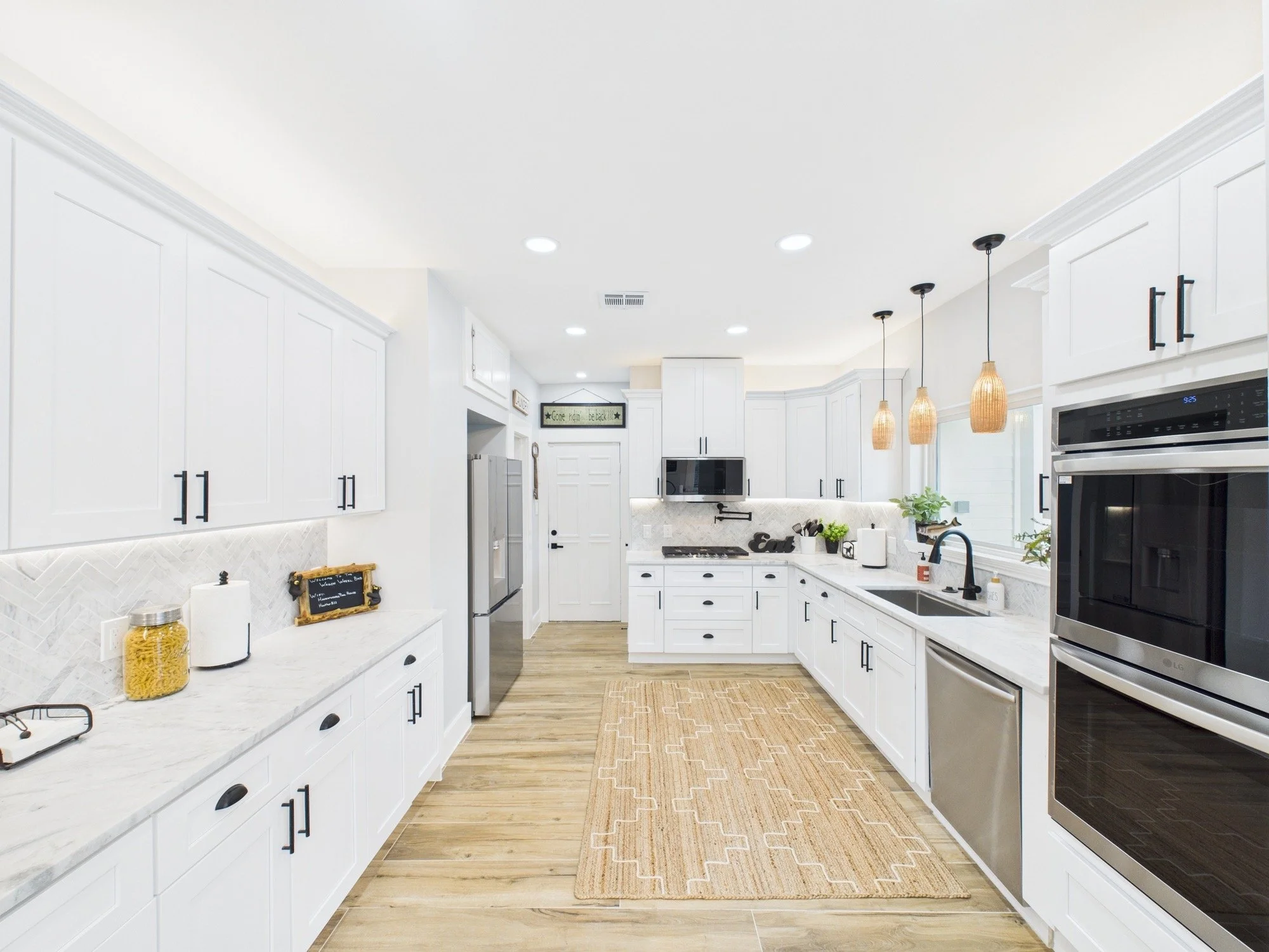 Modern white kitchen with hanging pendant lights, stainless steel appliances, and a beige patterned rug on wood flooring.