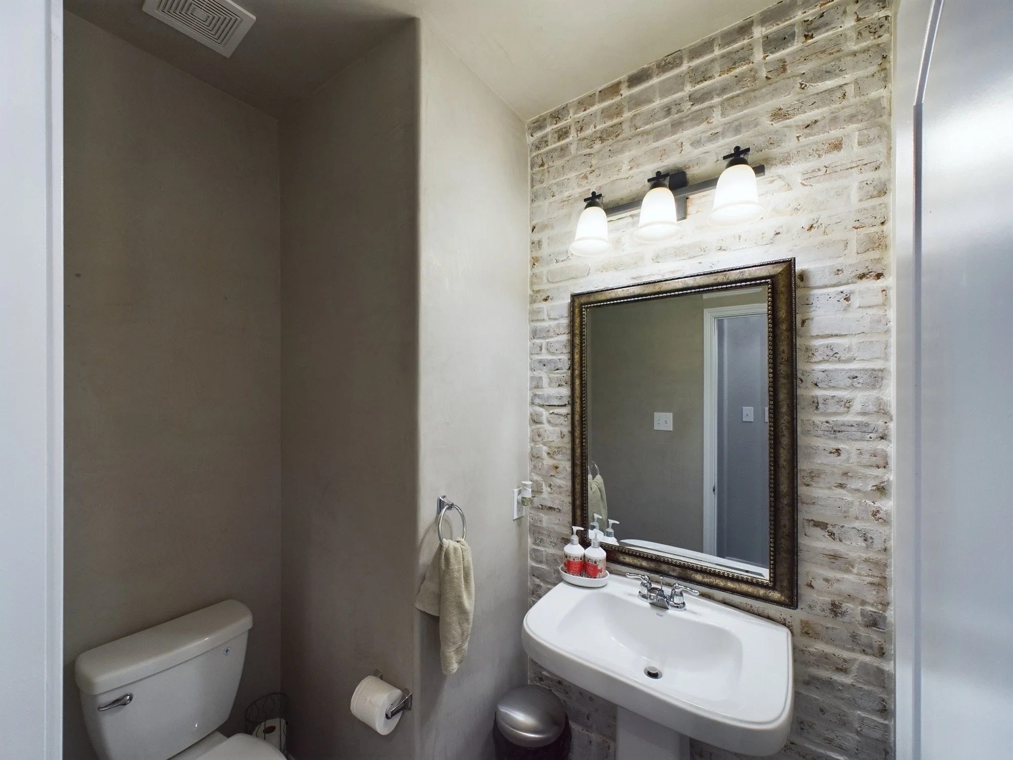 Small bathroom with a brick accent wall, a mirror above a sink, a light fixture with three bulbs, a toilet, and a towel hanging on a hook.