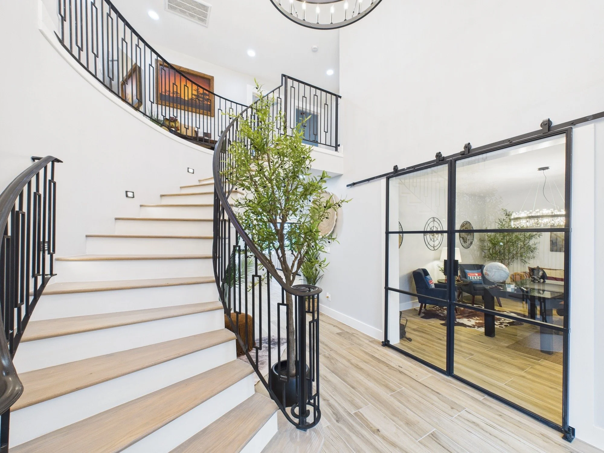 Interior view of a modern home featuring a staircase with wooden steps and black metal railing, a large potted plant at the base of the stairs, and a room separated by a glass partition behind sliding doors. The space has minimalist decor with white 