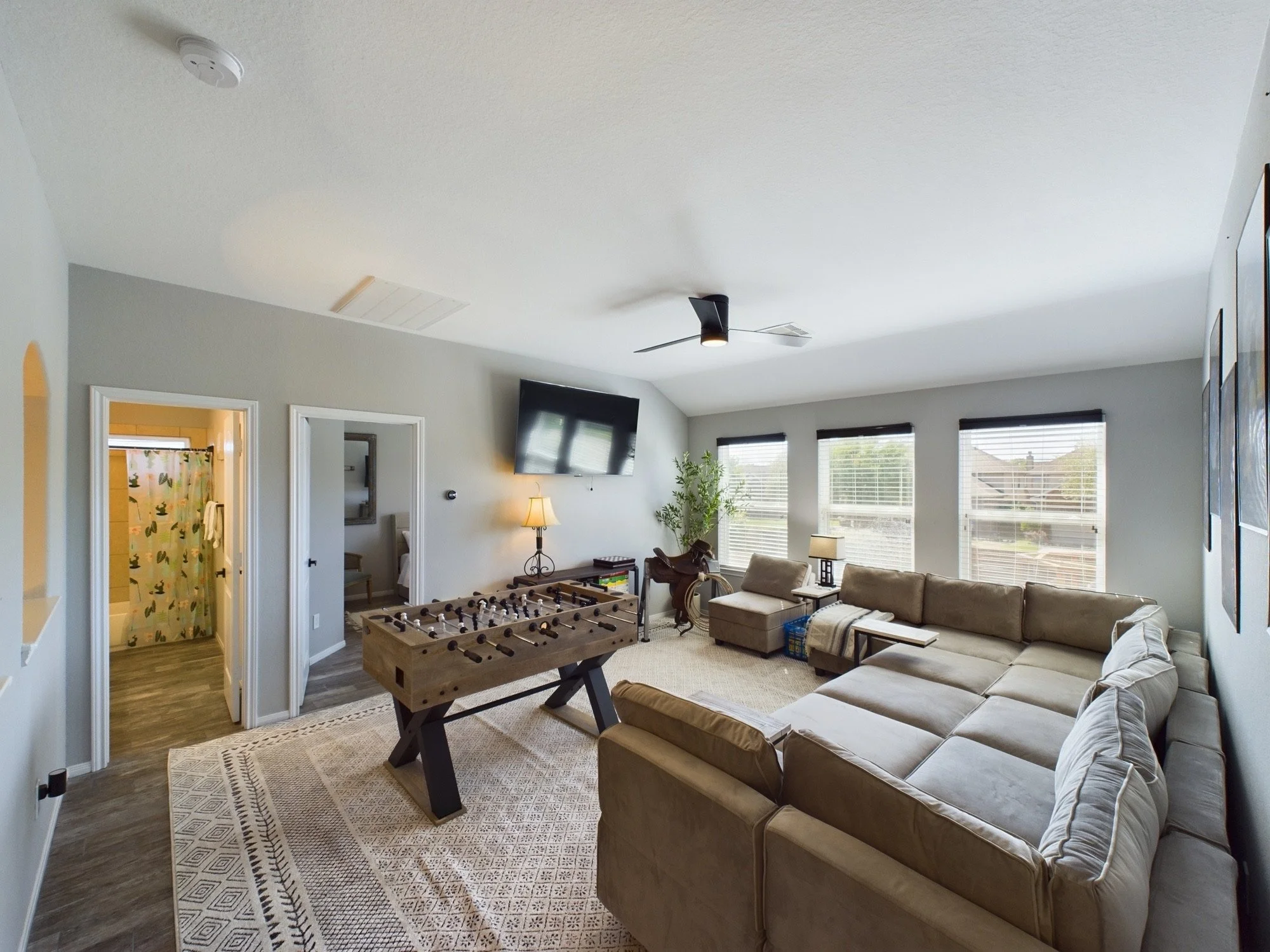 Living room with beige sofas, a foosball table, large windows with blinds, a flat-screen TV mounted on the wall, a standing lamp, and a plant. In the adjoining room, a bathroom with a shower curtain featuring butterfly prints is visible.