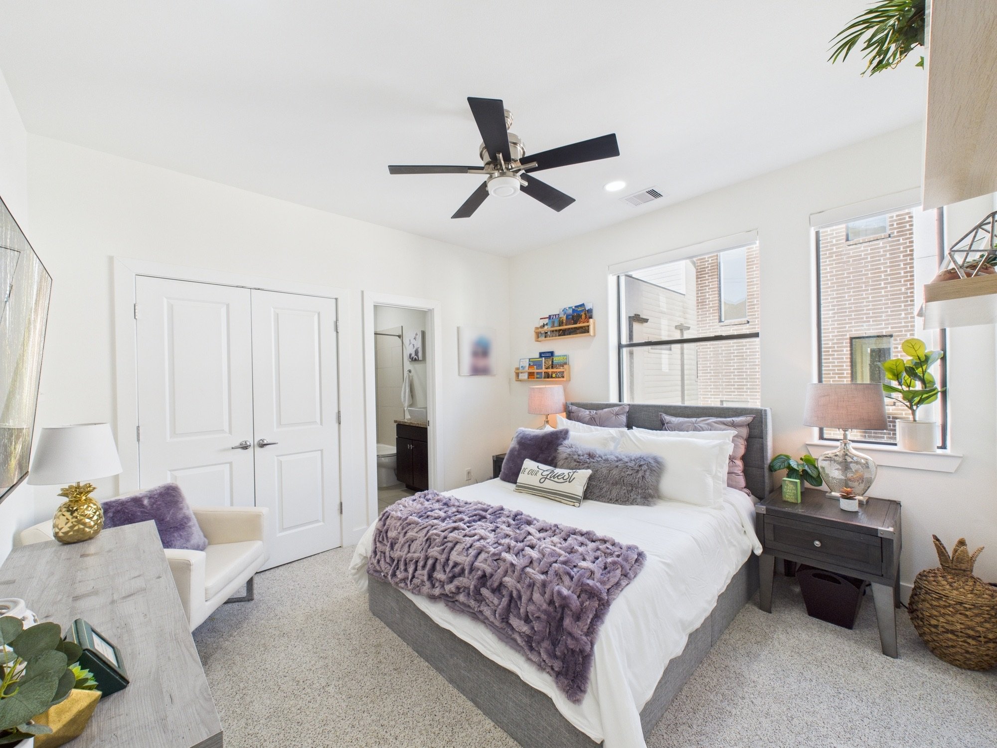 Bright bedroom with white walls and large windows, a bed with purple and gray throw pillows and a plush purple blanket, a black nightstand, a lamp, plants, and decor, with art and shelves on the wall.