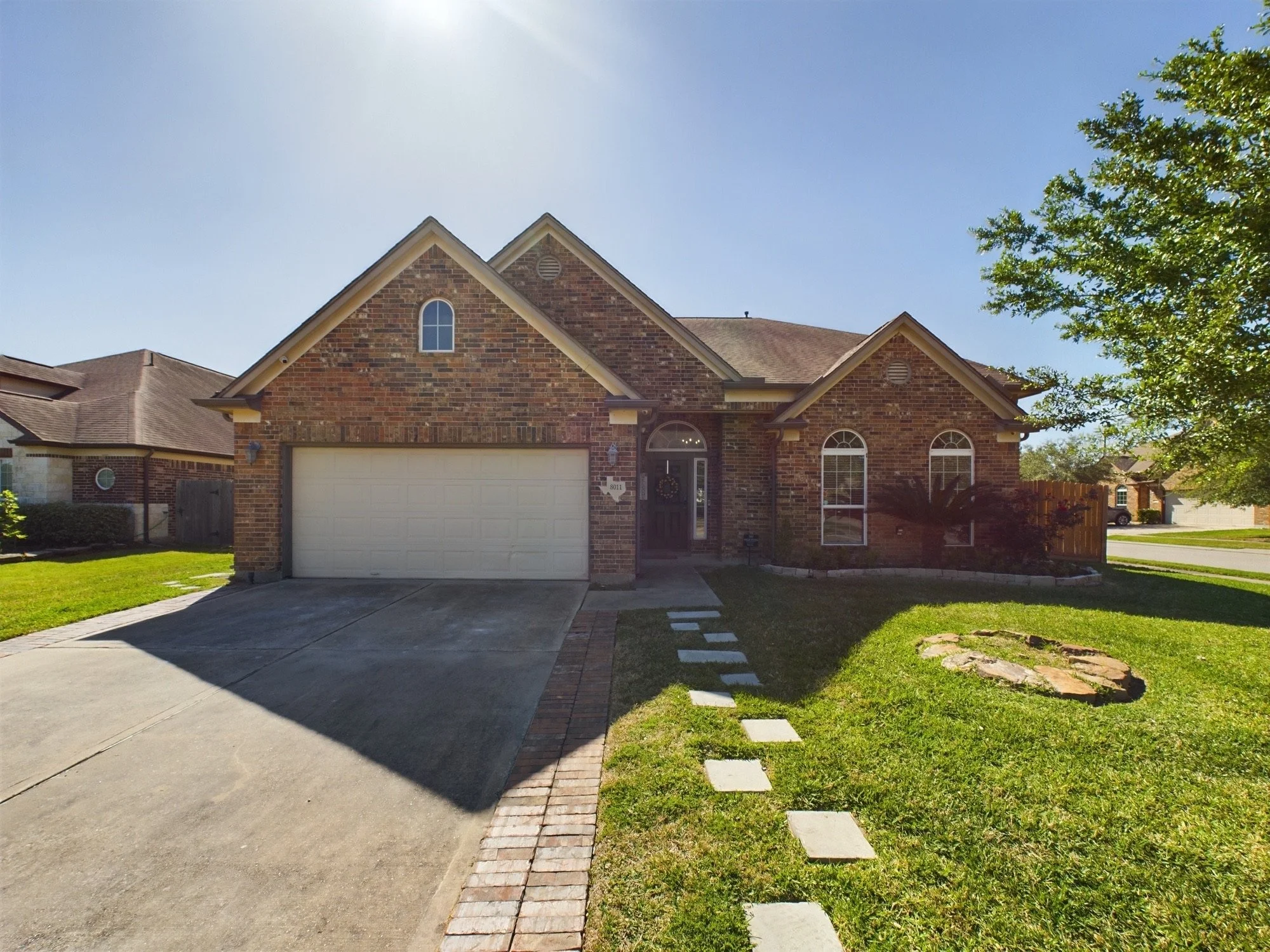 A two-story brick house with a white garage door, arched windows, and a front door decorated with a wreath. The house has a lawn on each side, a concrete driveway, and a walkway with square stepping stones. There is a tree on the right and neighborin