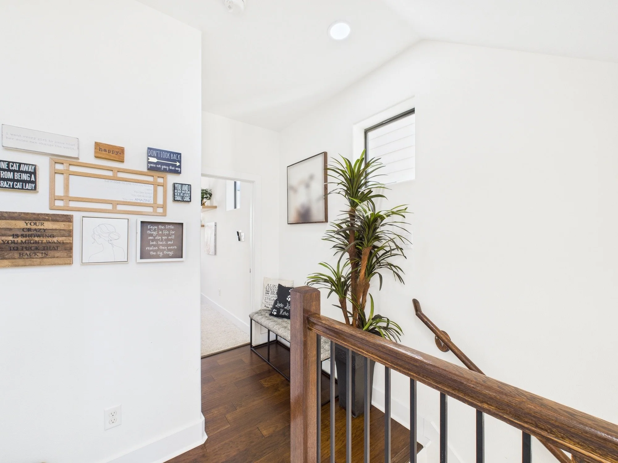 Interior view of a staircase landing with a white wall decorated with framed artwork and quotes, a large potted plant, a bench with pillows, and a window letting in natural light.