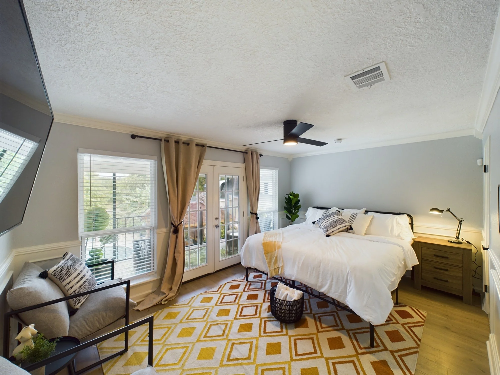 A bright bedroom with a white bed, patterned pillows, a wooden nightstand, black lamps, and large windows with blinds and beige curtains, overlooking a backyard with trees and a deck.