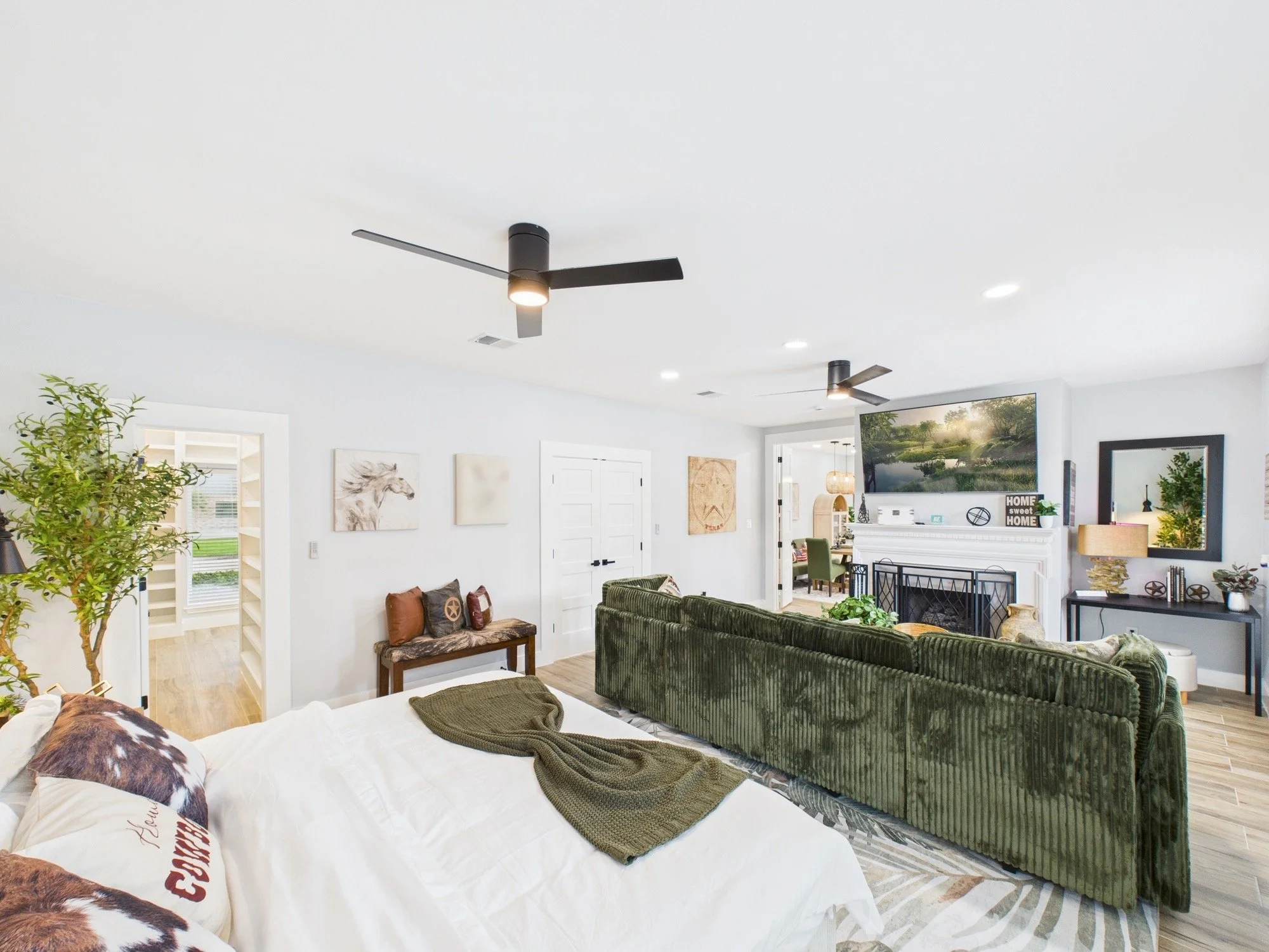 Living room with white walls, ceiling fans, wall art, a large green velvet sofa, a white fireplace, and a TV mounted above it. There are lights on the ceiling and houseplants in the room.