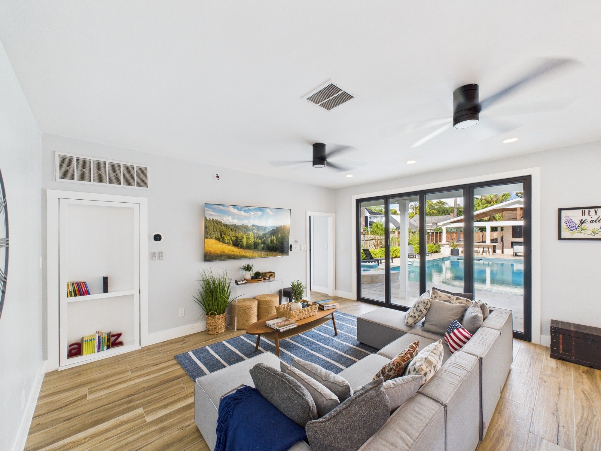 A modern living room with a gray sofa, blue and patterned pillows, a wooden coffee table, and a striped rug. A TV displays a landscape and is mounted on a white wall. Large glass sliding doors lead to an outdoor pool area with outdoor furniture and g
