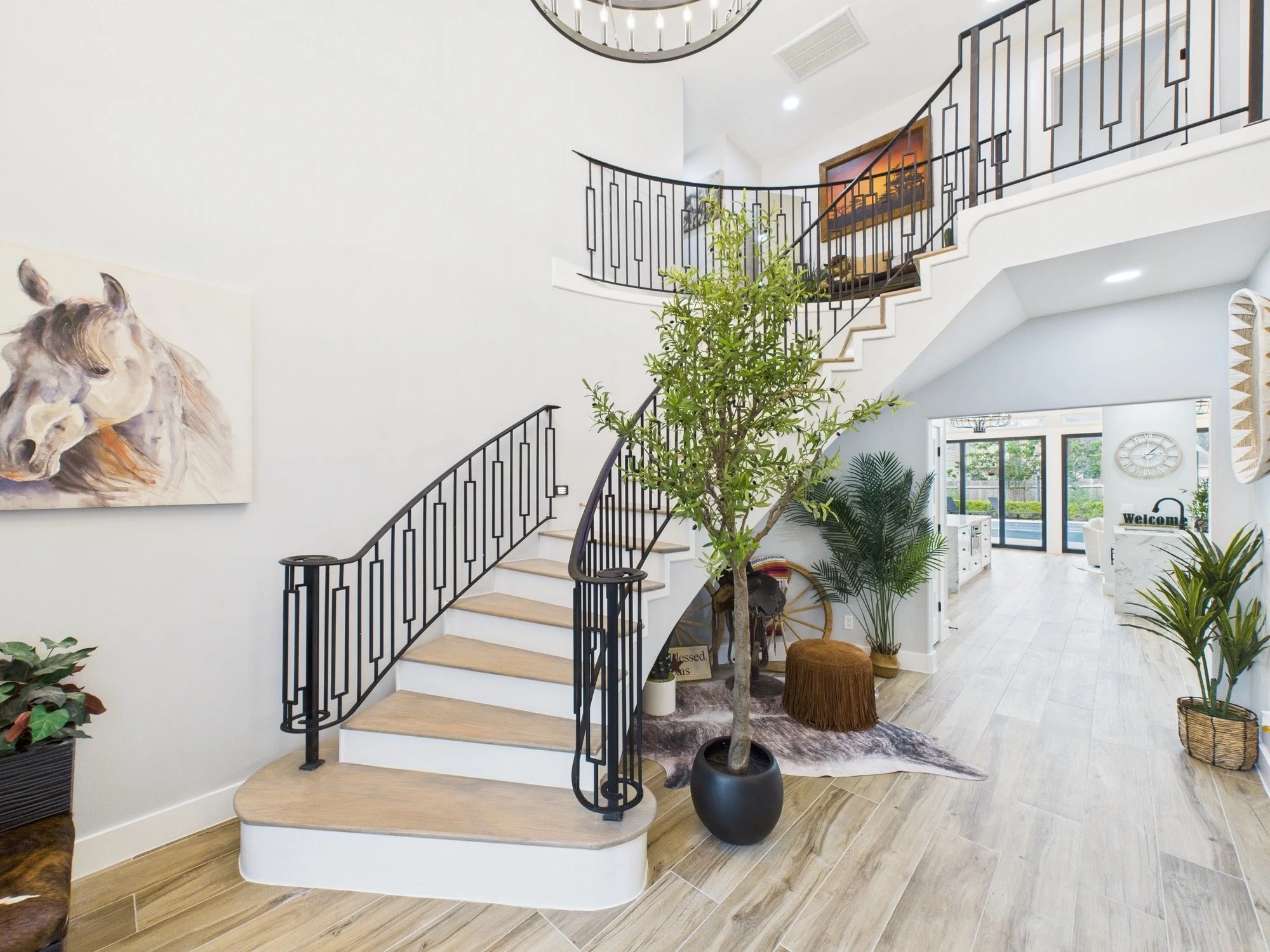 Interior of a modern home with a curved staircase featuring black railings and wooden steps, decorated with green plants and artwork, bright open living space with large windows and a chandelier.