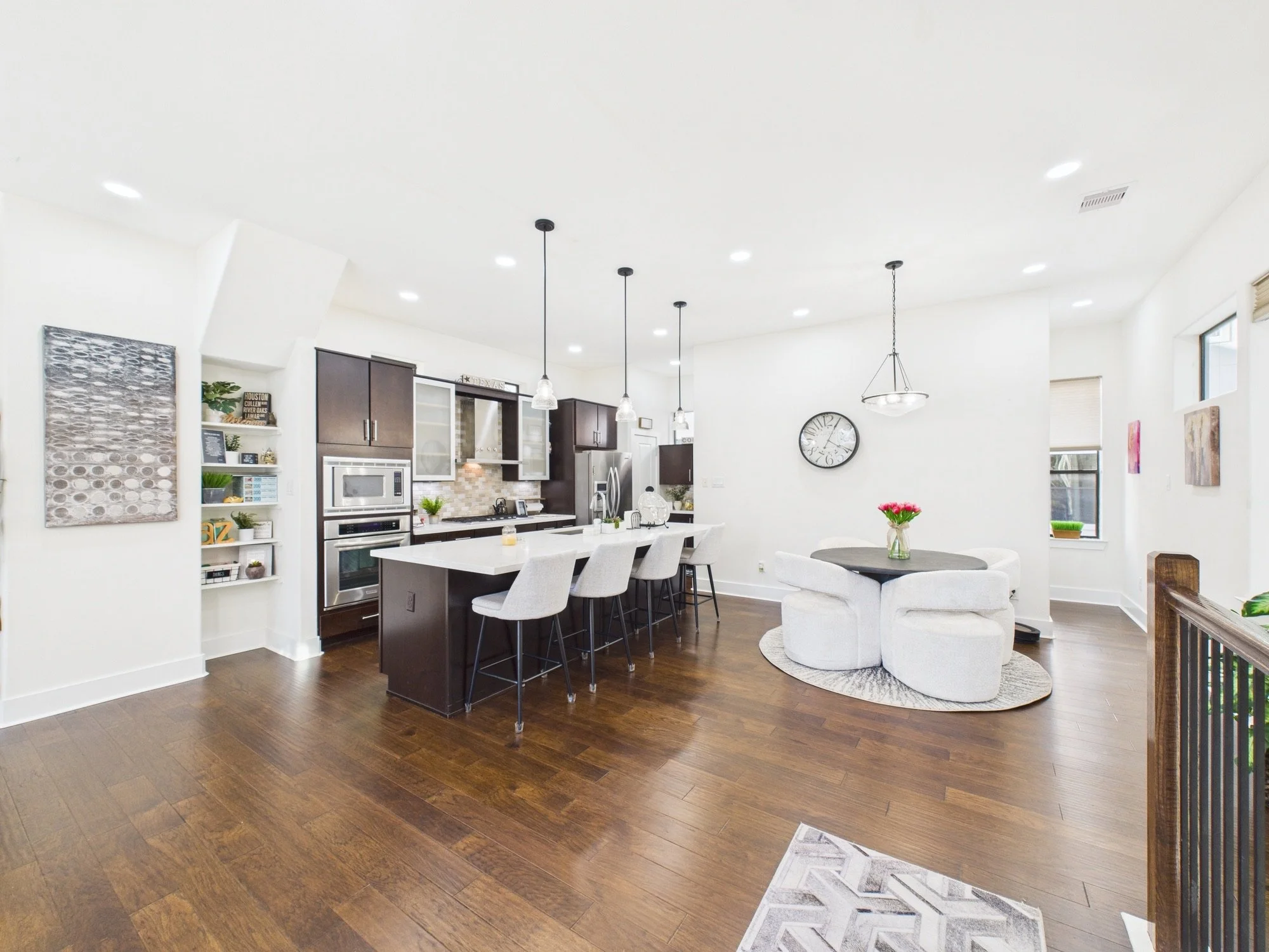 Open-concept living area with dark hardwood flooring, white walls, modern kitchen with dark cabinets, island, and stainless steel appliances, and a dining space with white chairs and a round table with a vase of pink flowers.