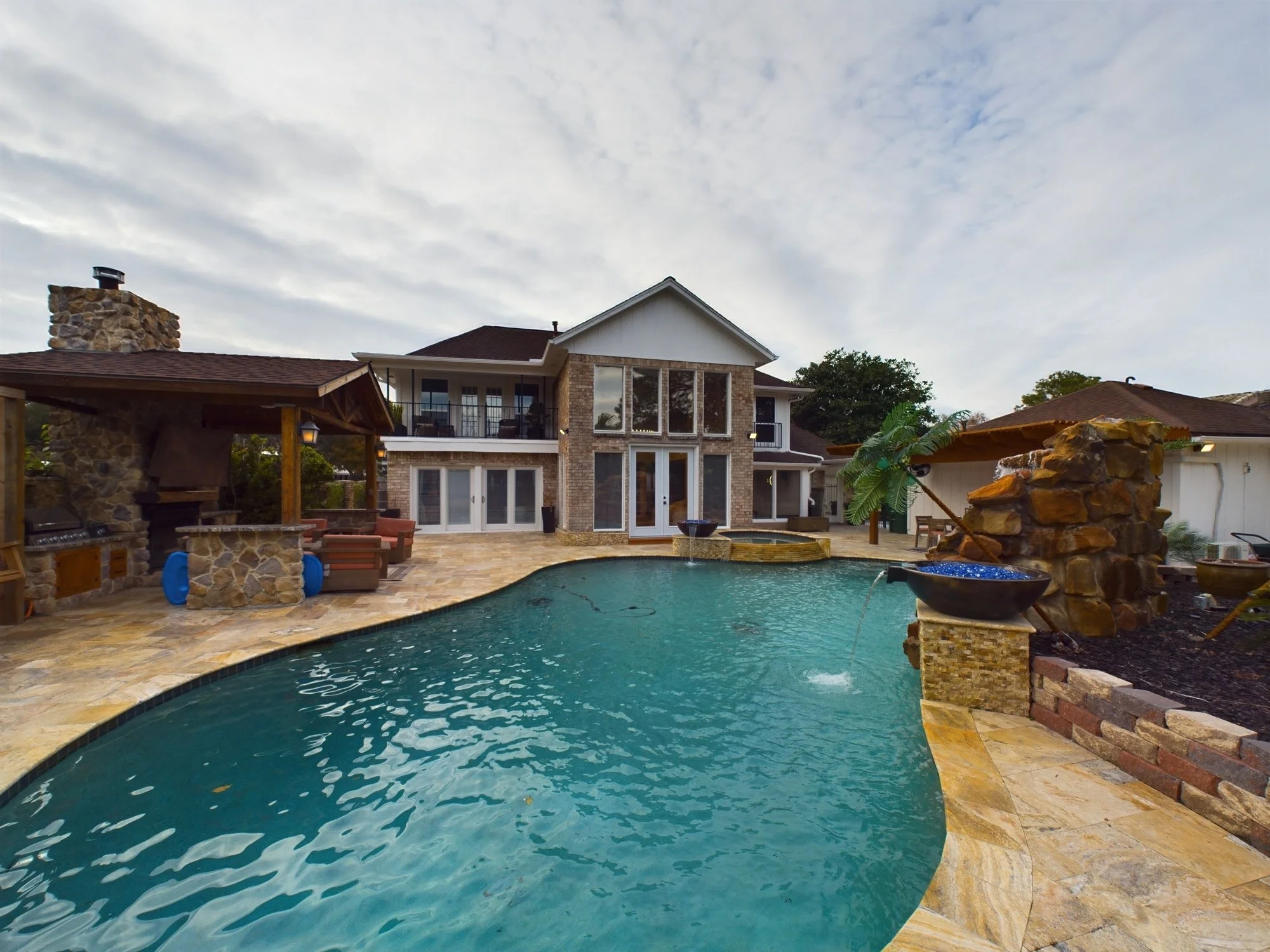 A backyard with a swimming pool, stone patio, outdoor seating, and a house with multiple levels and large windows at dusk.