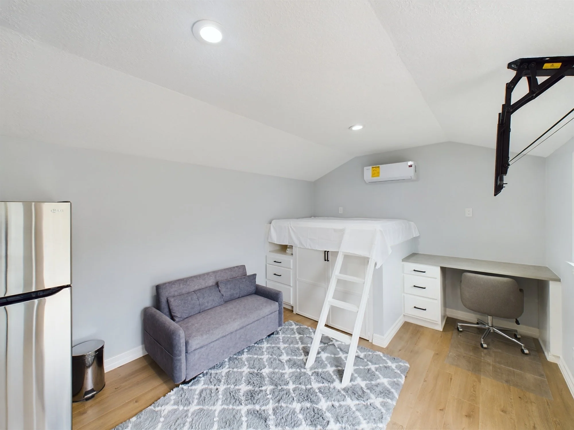 Living space with white walls and ceiling, two recessed lights, a gray sofa, a white dresser, a ladder, a mounted TV lift, a mini fridge, and hardwood flooring with a rug.