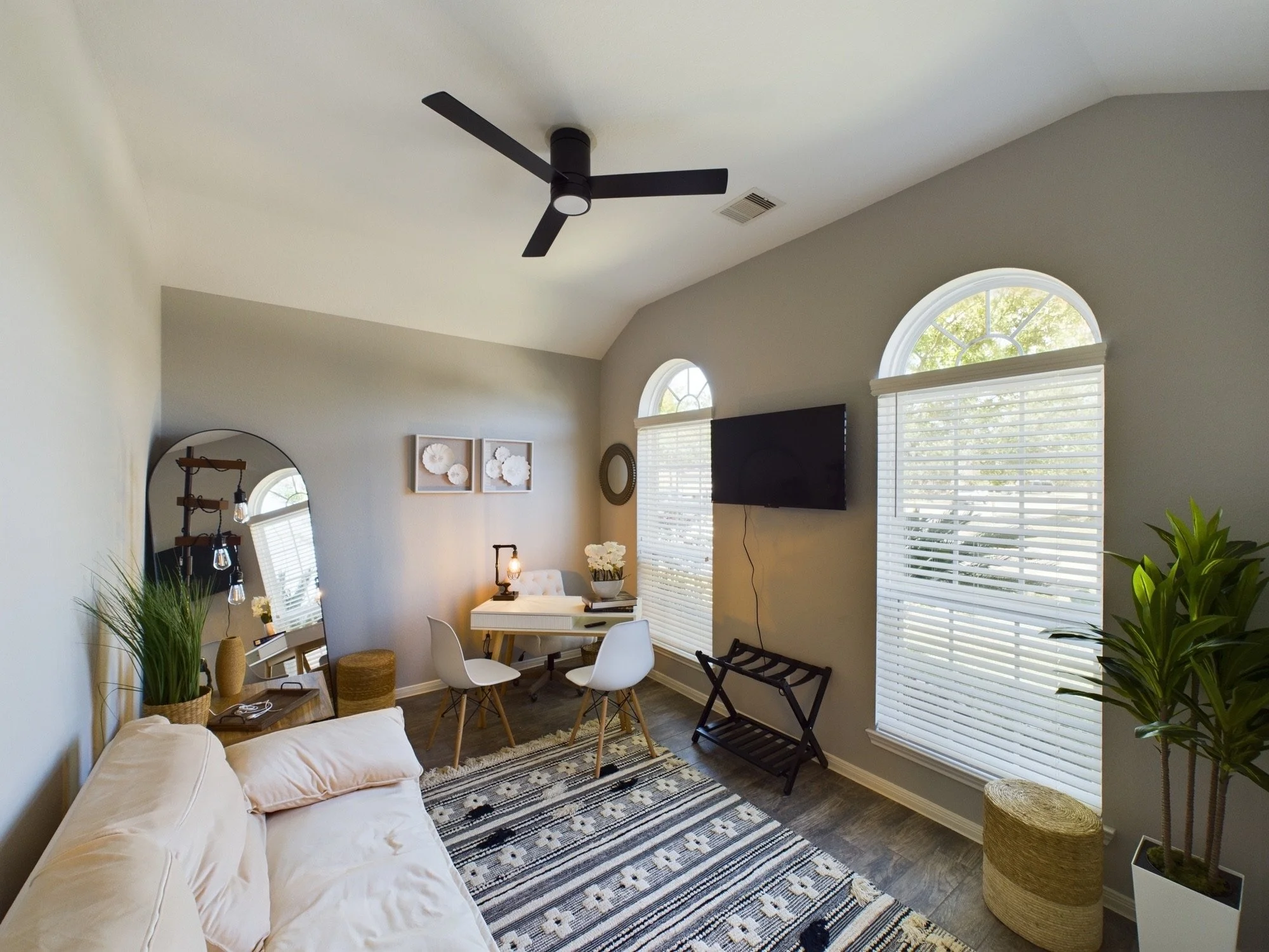 Living room with a white couch, a patterned rug, a small dining area with three white chairs, large arched windows with blinds, a wall-mounted TV, and various decorative items including plants and artwork.