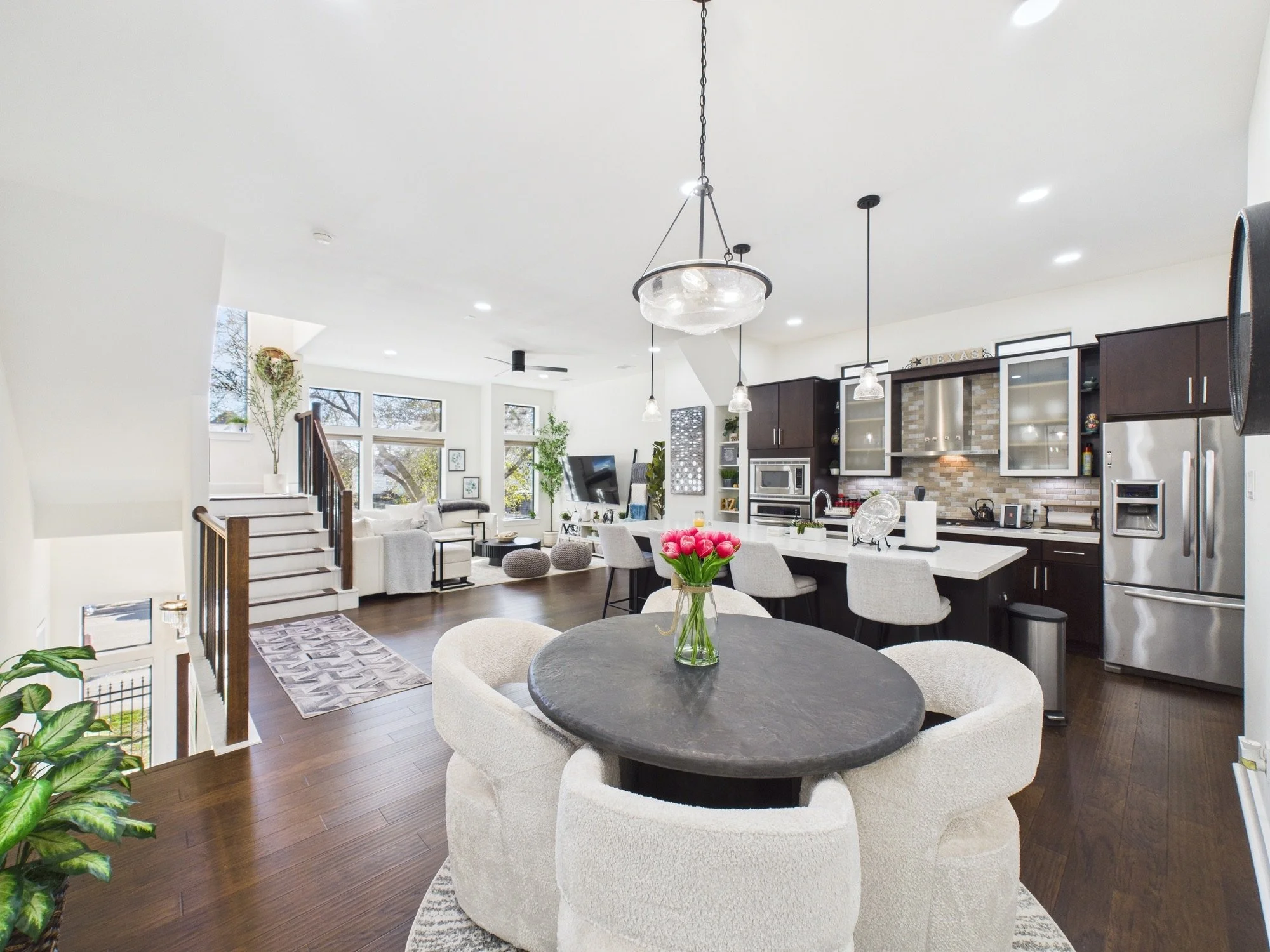 Open-concept kitchen and living area with white walls, dark wood cabinets, stainless steel appliances, pendant lighting, dining table with chairs, and a tv viewable from the dining table.