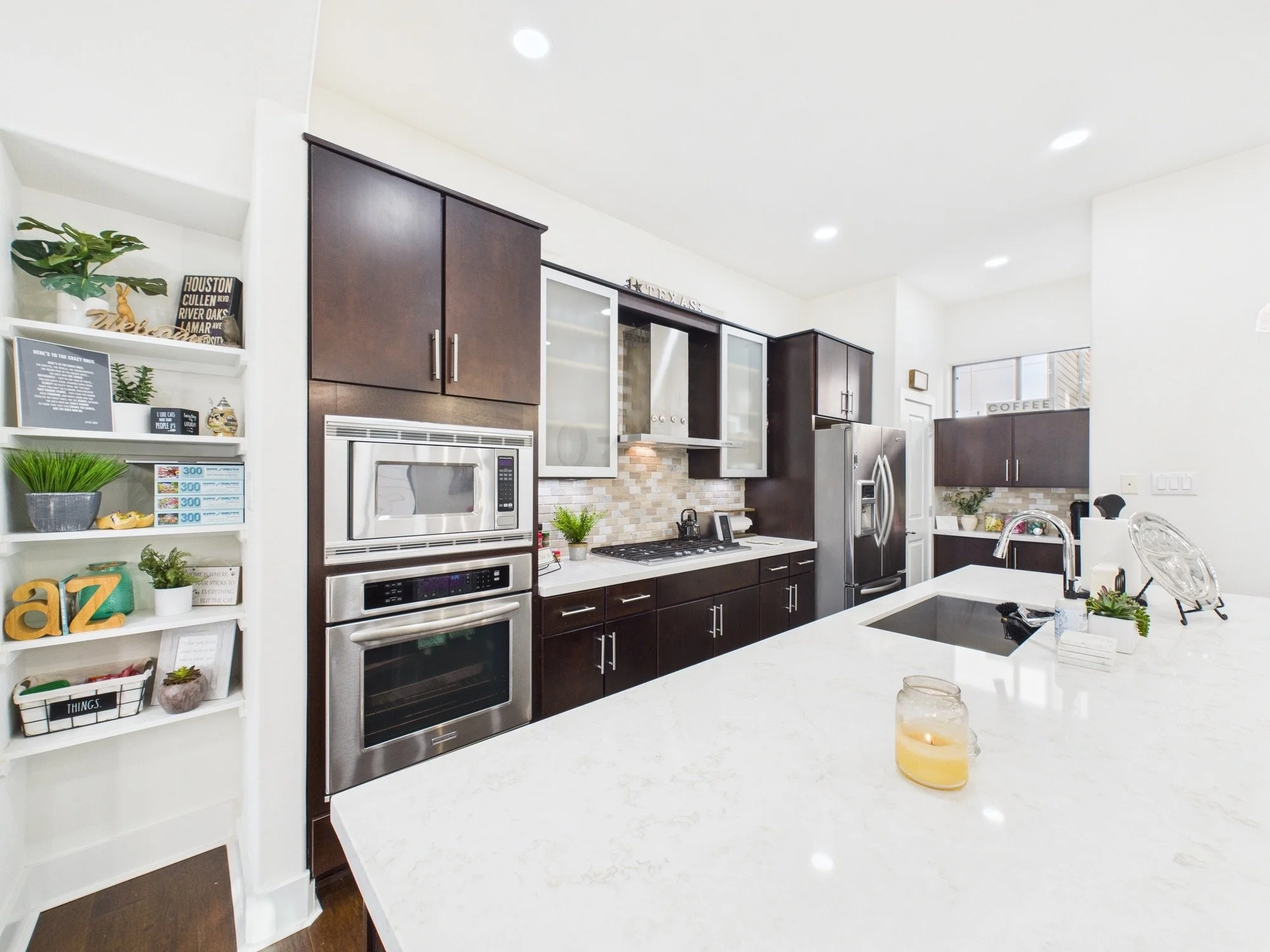 Modern kitchen with dark cabinets, stainless steel oven and refrigerator, white countertops, decorative items, plants, and a candle on the island.