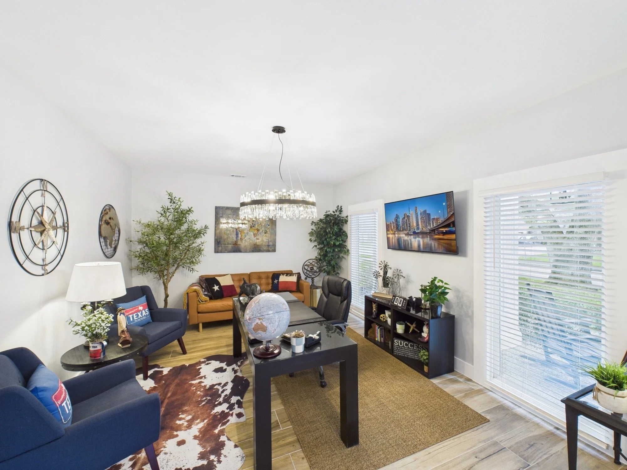 Living room with blue and tan furniture, world globe, plants, wall decor, window blinds, and a TV on the wall showing cityscape