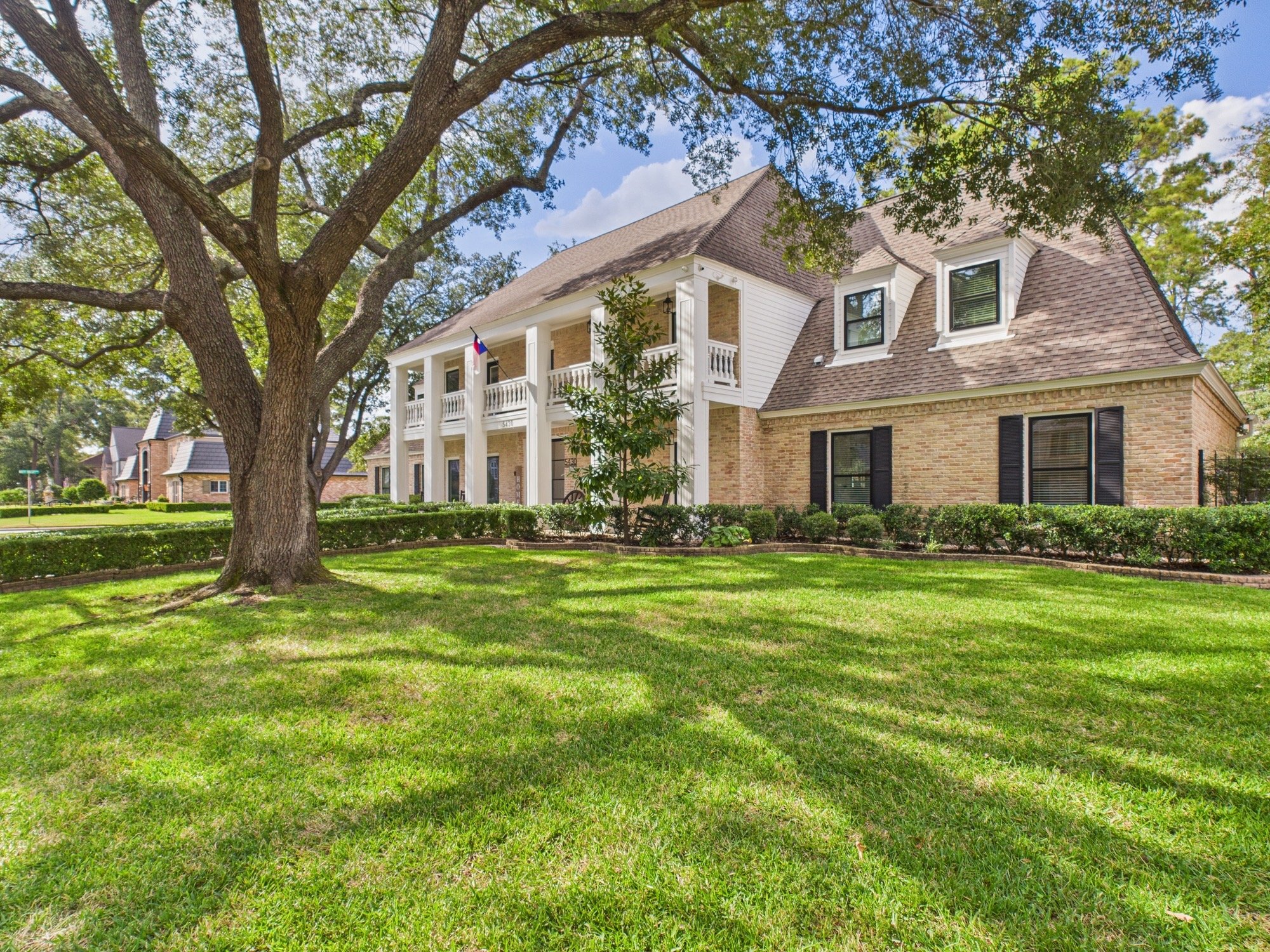 A large residential house with brick and white siding, a gray shingled roof, a front porch with columns, and a Texas flag hanging from a pole. There is a large tree in the front yard with a lush green lawn surrounding it, and neighboring houses are v