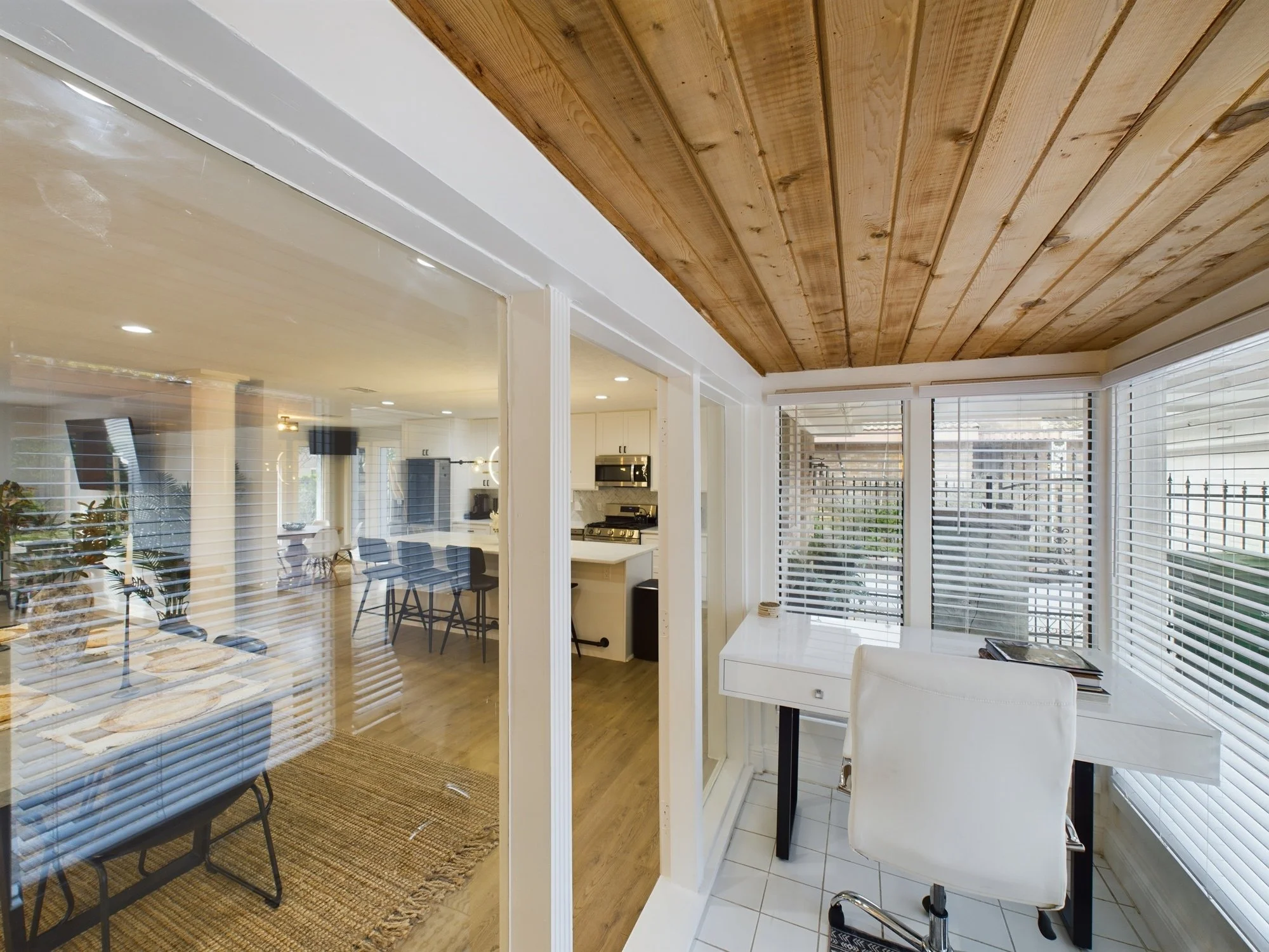 Living room with large windows, white walls, wooden ceiling, and a view into the kitchen and dining area with modern furniture.