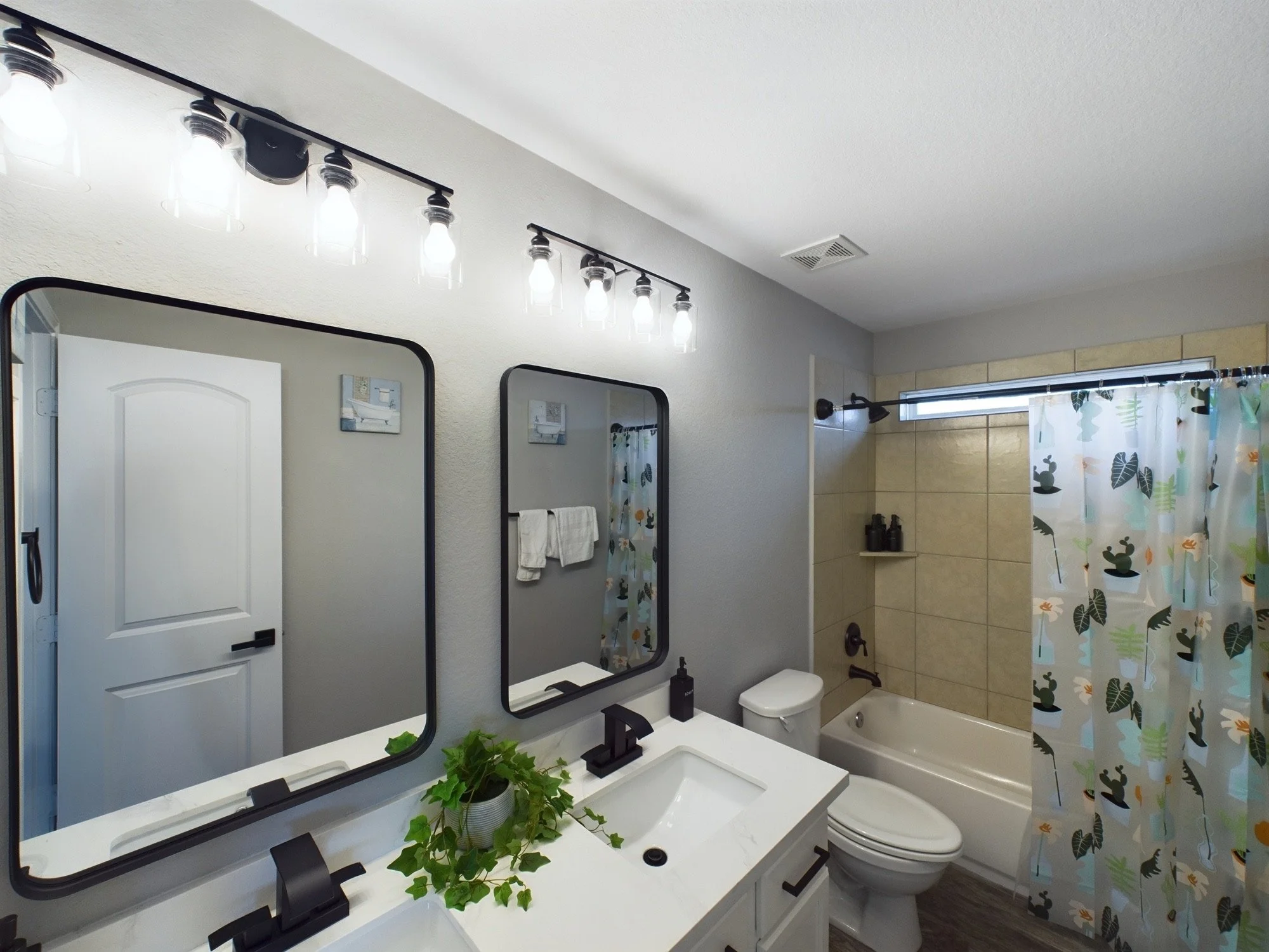 A bathroom with a white double vanity, black fixtures, and two rectangular mirrors. There is a small plant on the counter, a bathtub with a shower curtain with leaf patterns, a toilet, and a window with a curtain. Overhead, there is a light fixture w