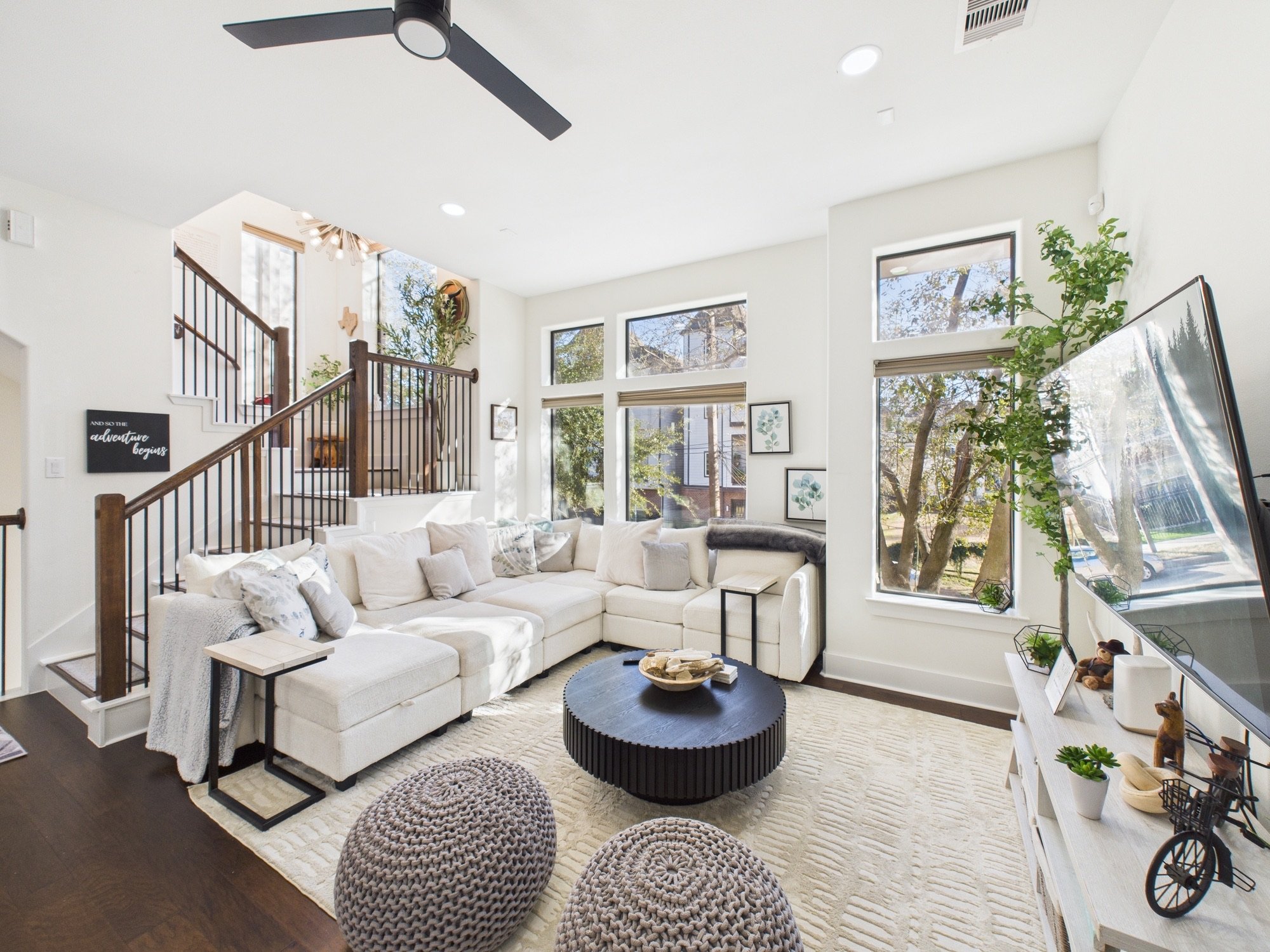 Bright living room with white sectional sofa, black coffee table, large windows, indoor plants, and personal decorative items.
