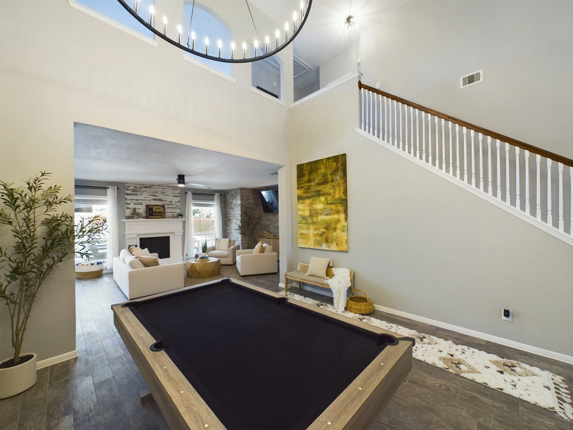 Living room with beige walls, white furniture, a fireplace, large windows with white curtains, a large abstract painting, a pool table, and a staircase with a wooden handrail