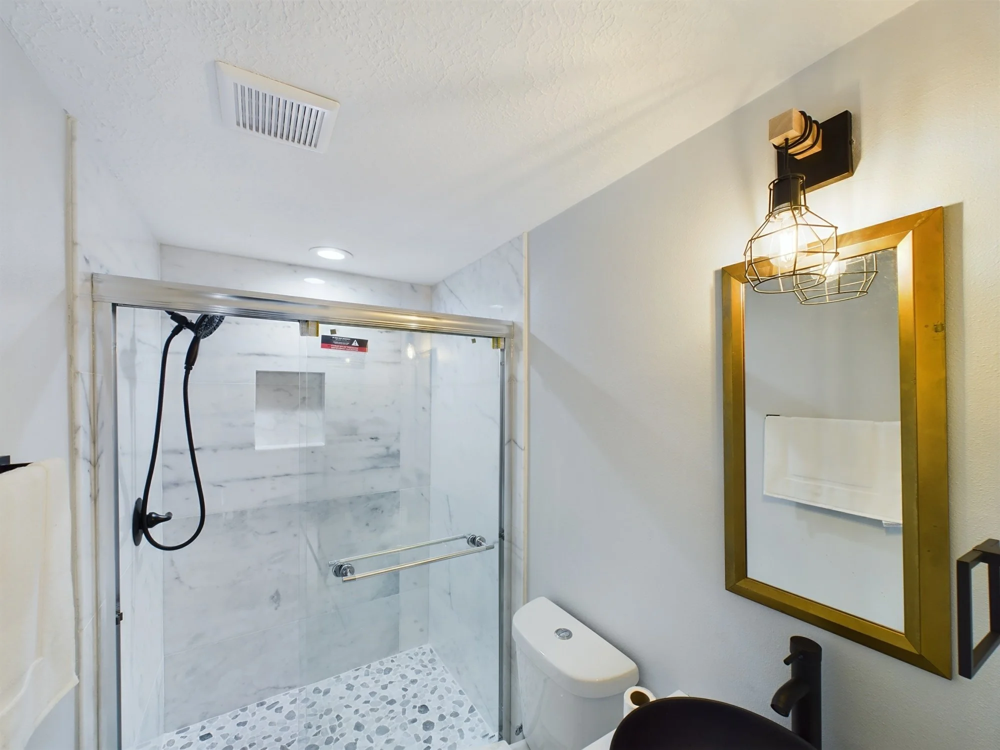 Bathroom with shower area featuring marble tiles, a glass door, a toilet, vanity mirror with gold frame, modern light fixture, and ceiling ventilation.