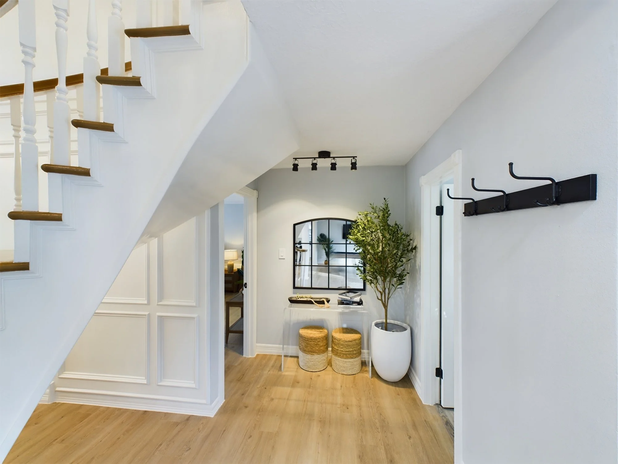 Entryway with wooden floors, white walls, a coat rack, a large potted plant, and a mirror in the distance.