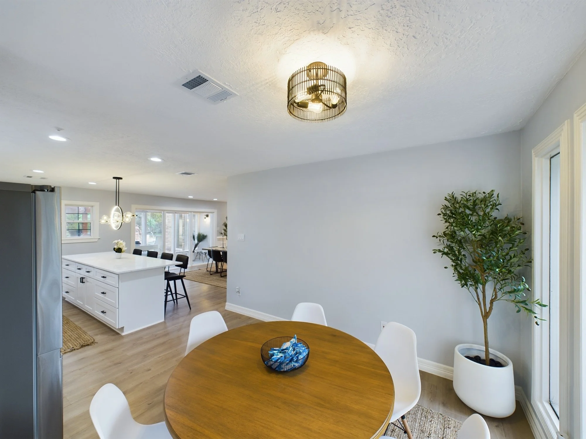 A bright, modern kitchen and dining area with a round wooden table, white chairs, a potted plant, and a decorative ceiling light fixture.
