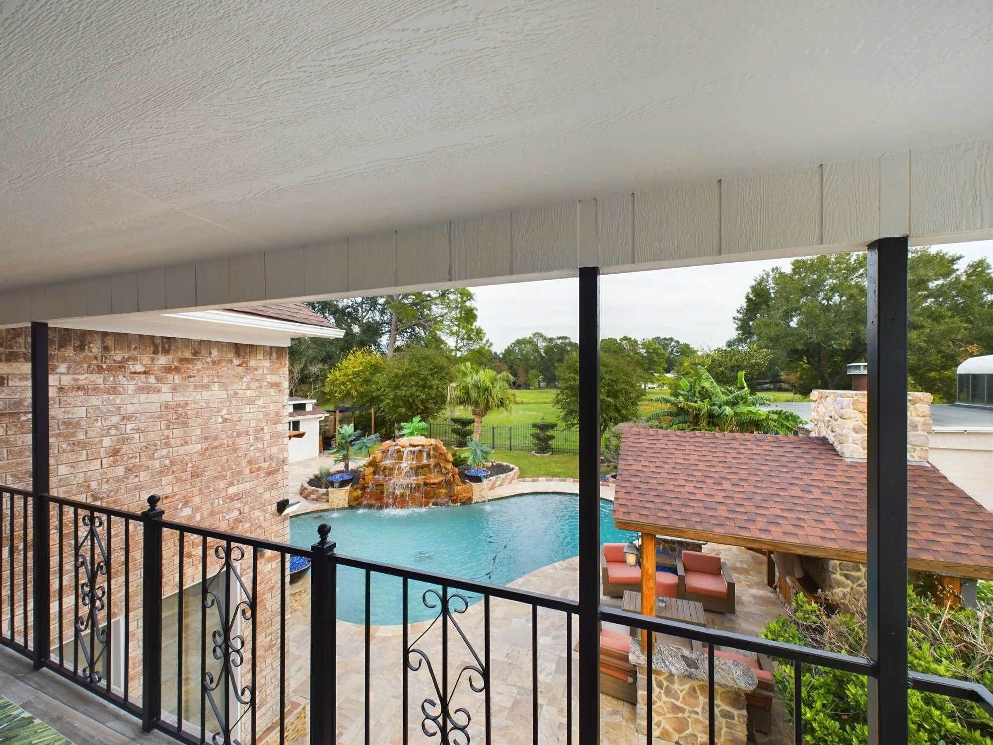 View of a backyard swimming pool with a waterfall feature, surrounded by a brick patio and lush greenery, seen from a covered patio with a black wrought-iron railing.