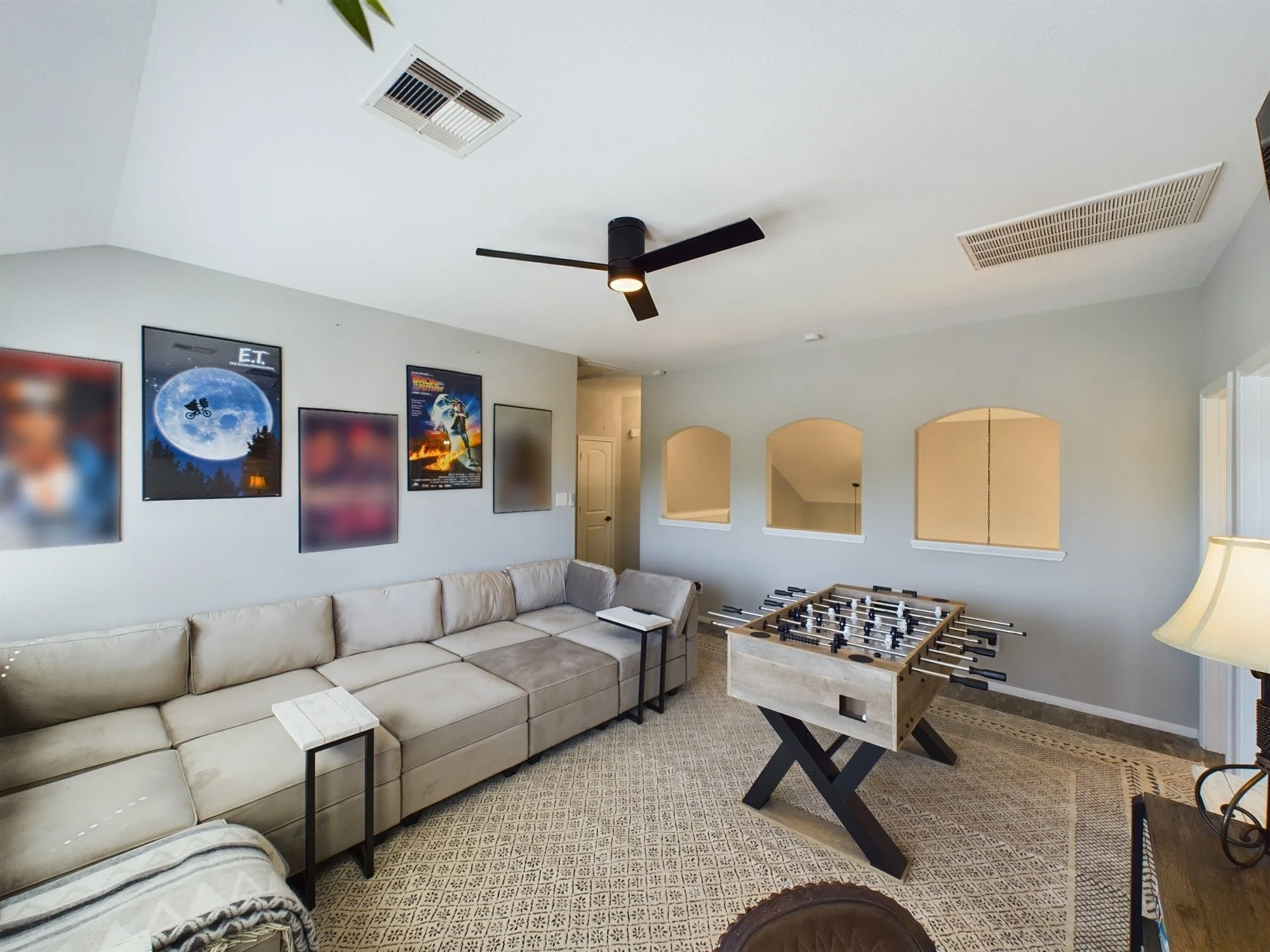 A living room with a beige sofa, a foosball table, a ceiling fan, wall art, a TV, and a patterned carpet.
