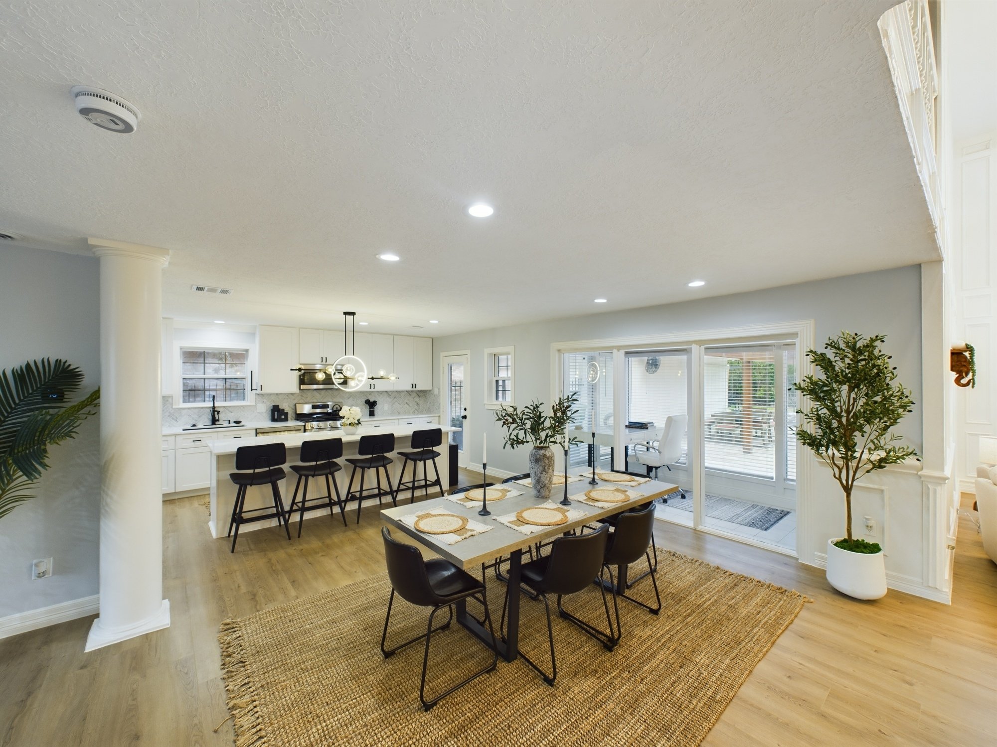 Open-concept living room and kitchen with hardwood floors, white cabinetry, large windows, a dining table with six chairs, and indoor plants.